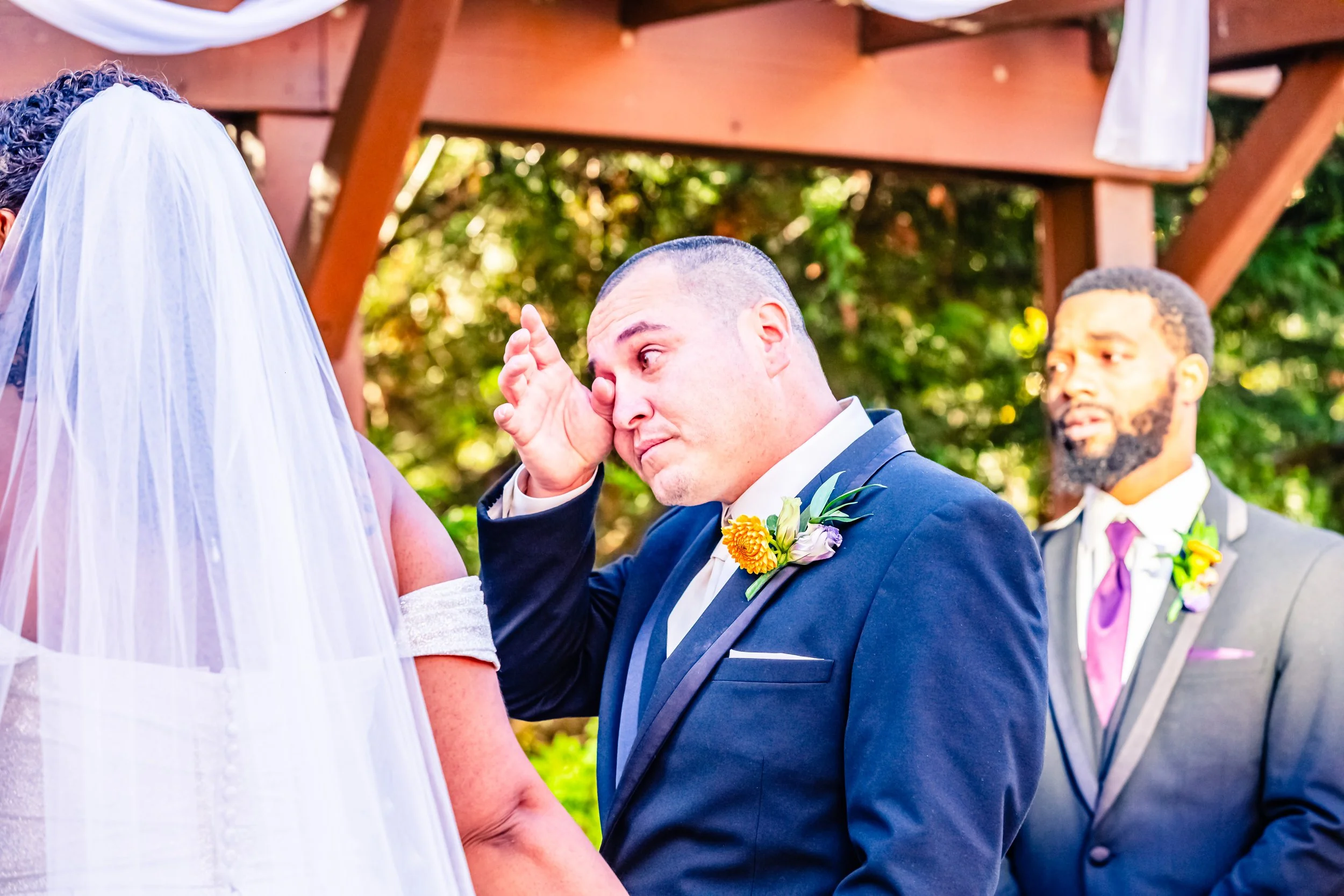 A groom wiping away tears during a wedding ceremony, standing next to a bride with a veil, with a groomsman in a suit in the background.
