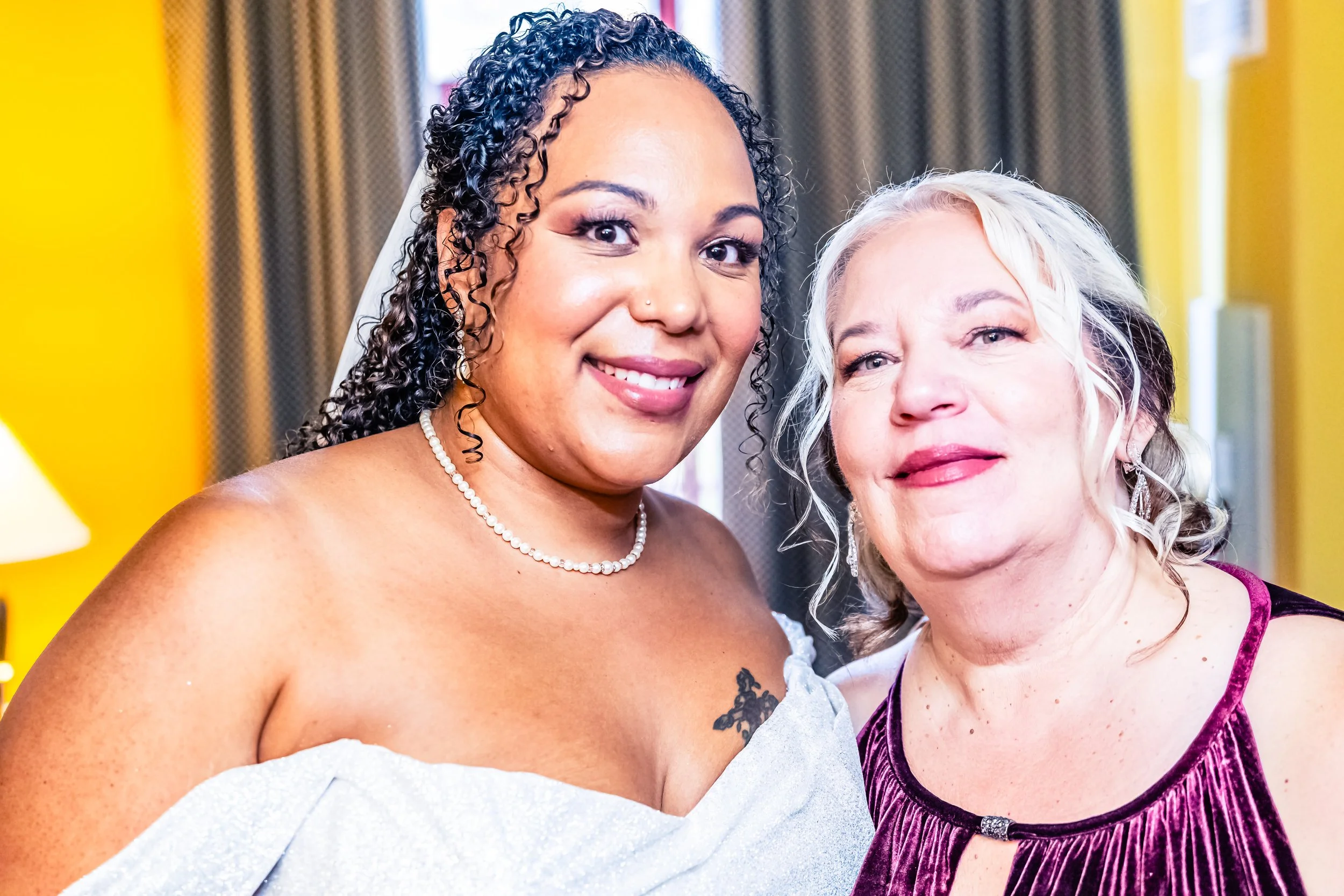 Two women, one with dark curly hair and the other with light blonde hair, smiling and standing close together in a brightly lit room.