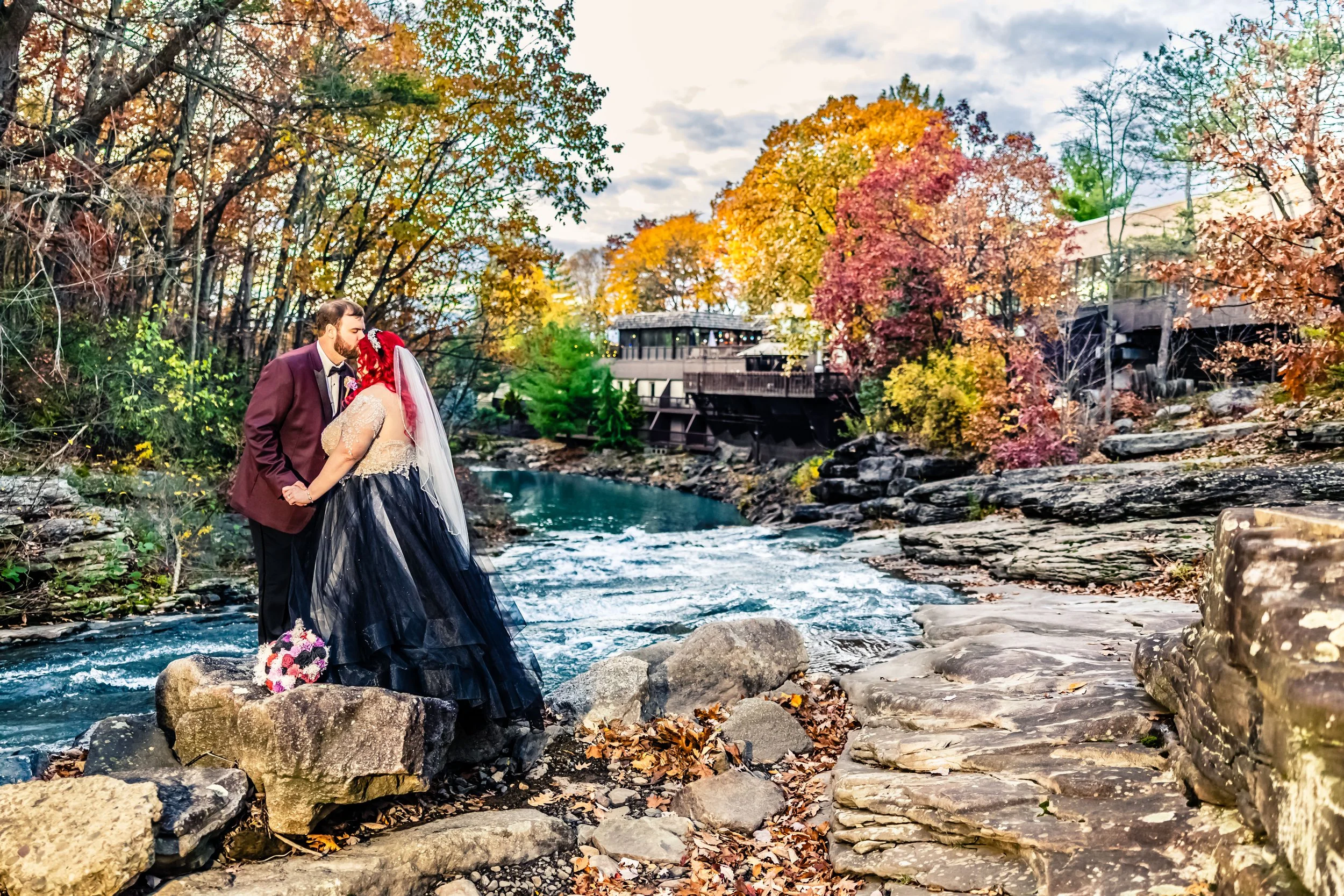 A newlywed couple kissing on rocks by a river with trees and a building in the background during autumn.