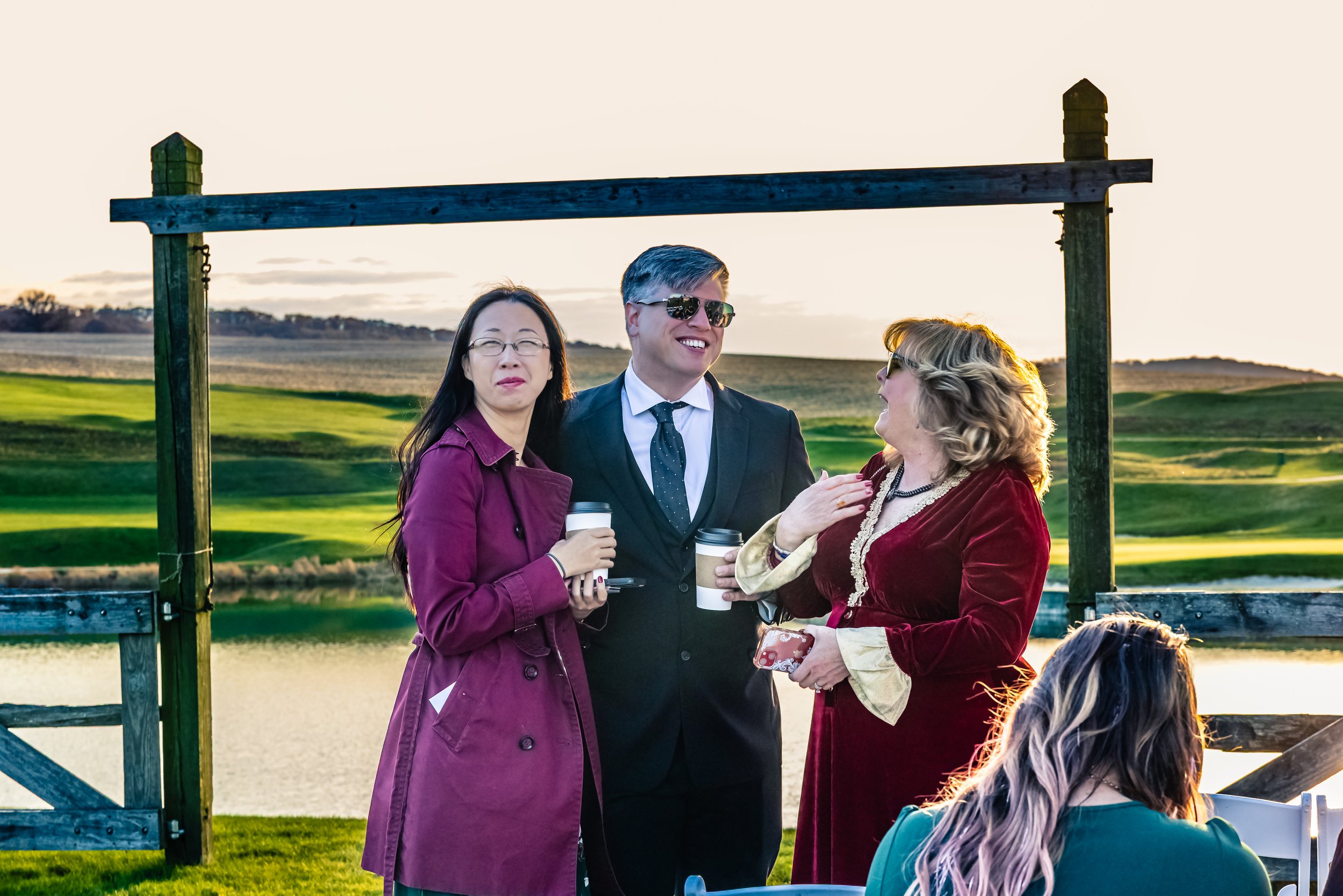 Three people standing by a lake at sunset, engaging in conversation, with one woman holding a paper coffee cup, a man in sunglasses smiling, and another woman talking animatedly. A woman with pink hair is seated in the foreground, facing away from th