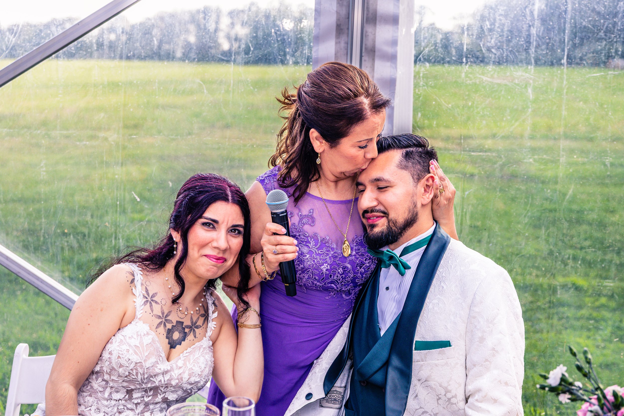 A woman in a purple dress is kissing a man in a white tuxedo on the forehead while holding a microphone, with a woman in a white lace dress sitting beside them smiling. They are inside a glass-walled venue with green grass outside.