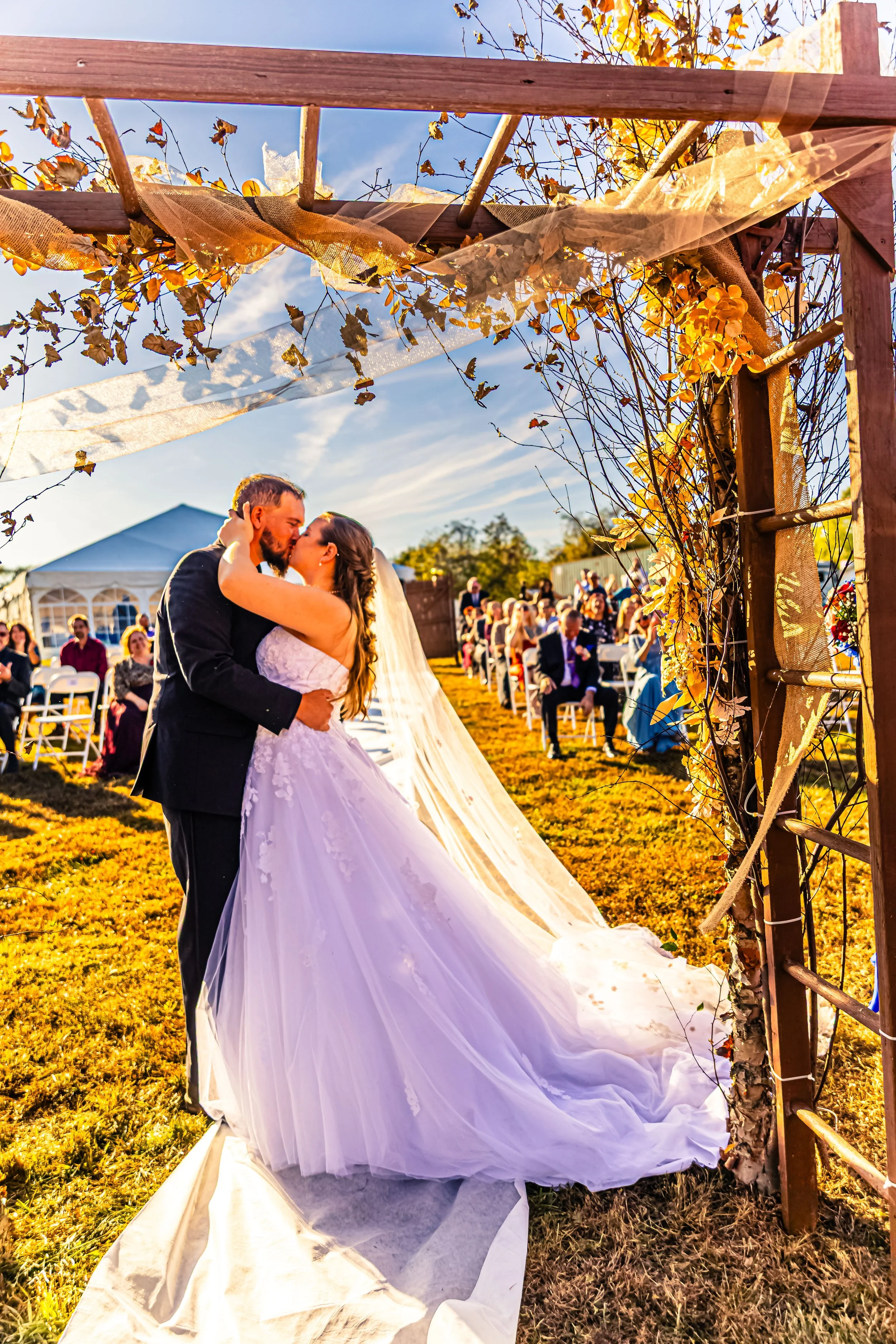 A bride and groom sharing a kiss during their outdoor wedding ceremony, with wedding guests seated in the background and a clear sky overhead.