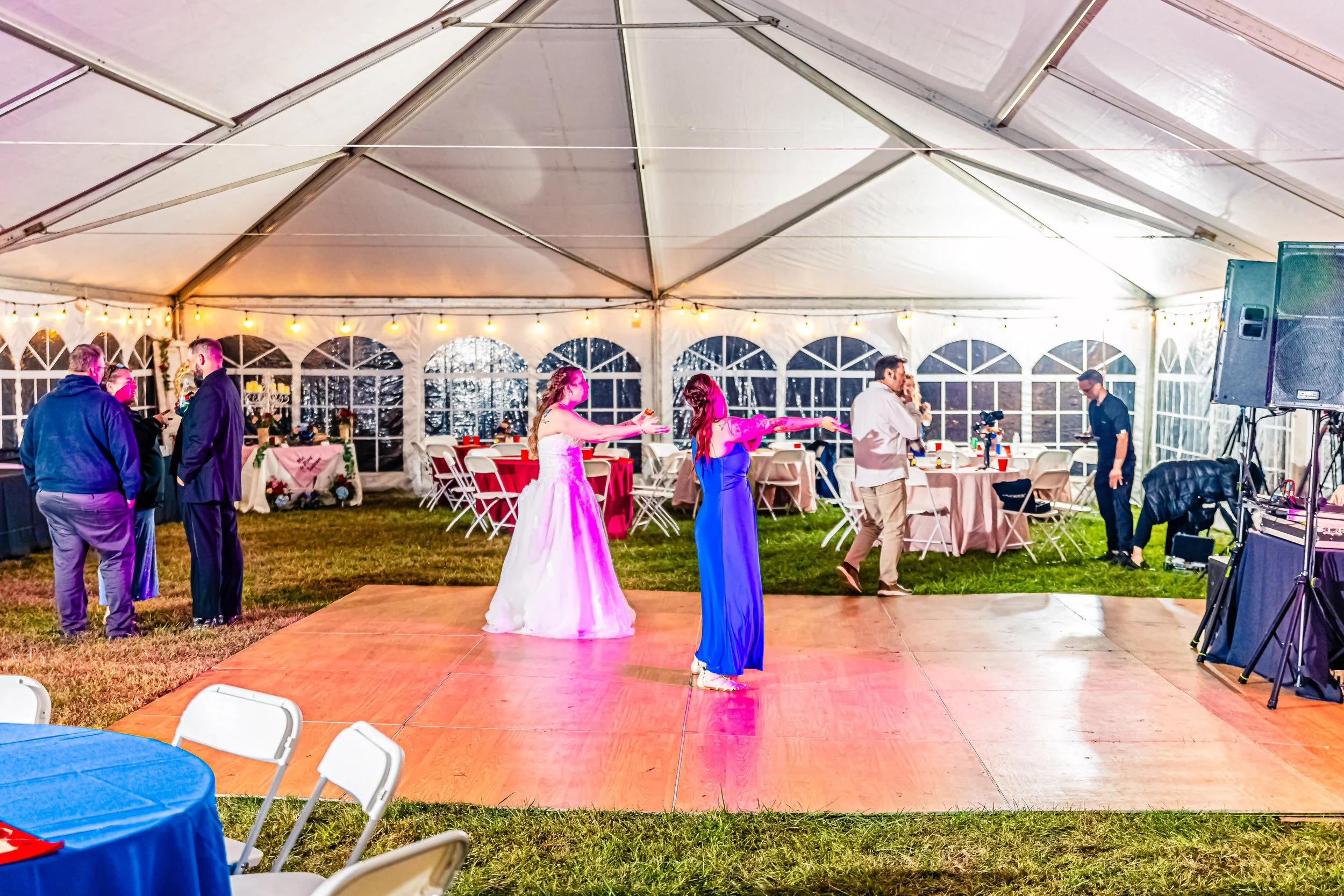 People dancing and socializing inside a large tent at a celebration with decorated tables and string lights.