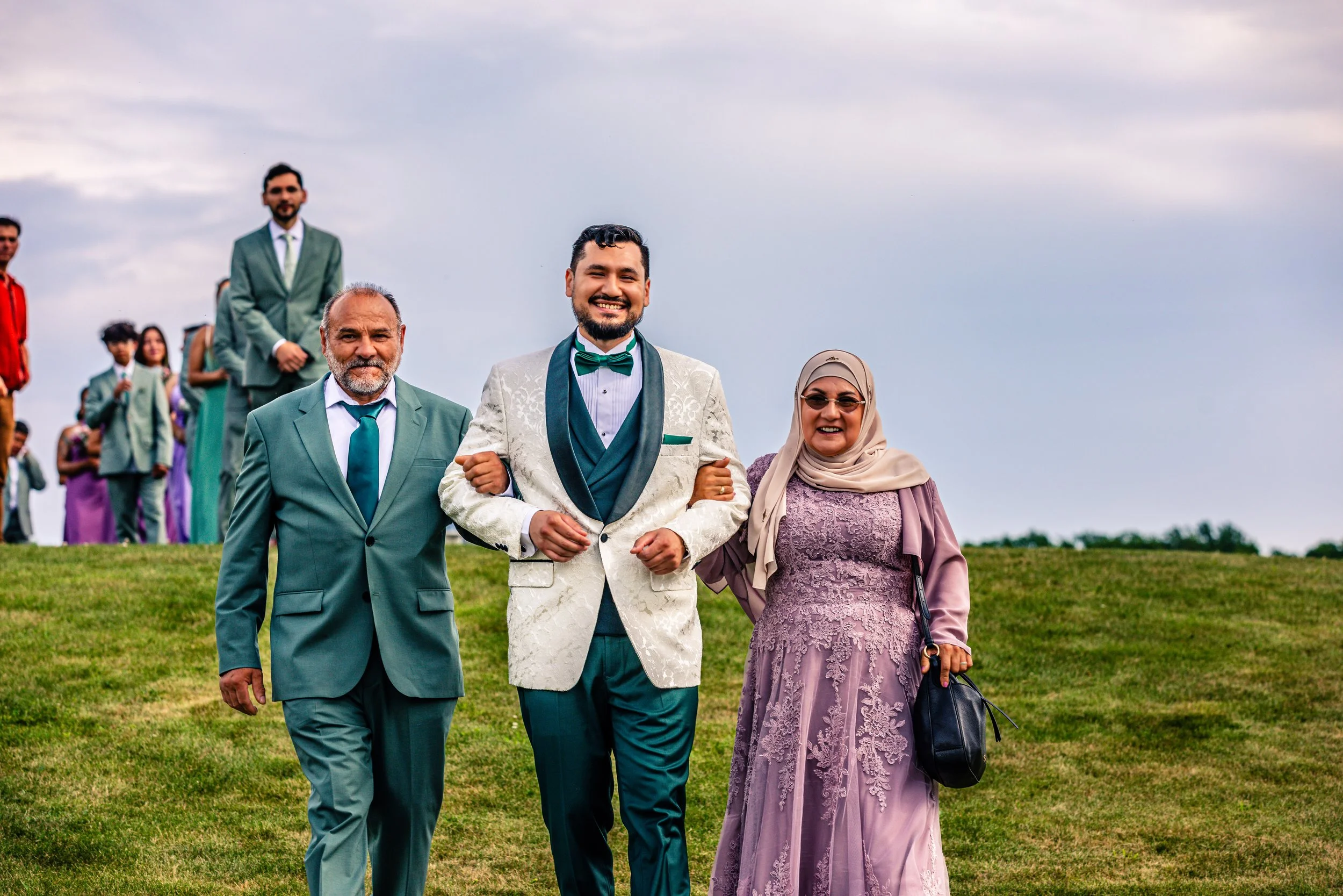 A man in a white tuxedo with a green bow tie, walking arm-in-arm with an older woman in a purple lace dress and hijab, during a wedding celebration on a grassy hill with wedding party members in the background.