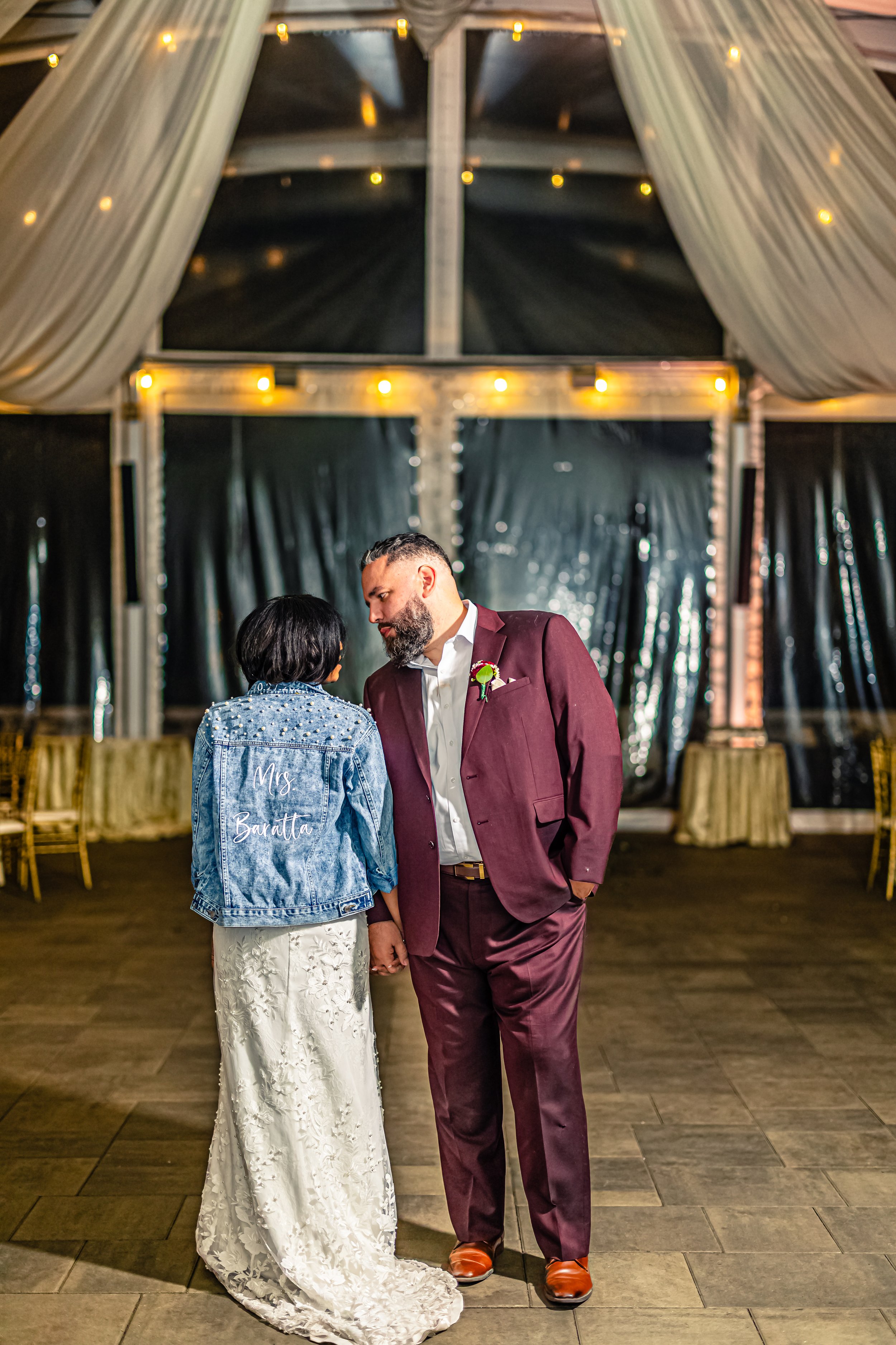 A bride and groom standing close together at their wedding reception inside a decorated tent, with the bride wearing a white lace dress and a denim jacket with 'Mrs. Banatta' embroidered on the back, and the groom in a burgundy suit with a boutonnier