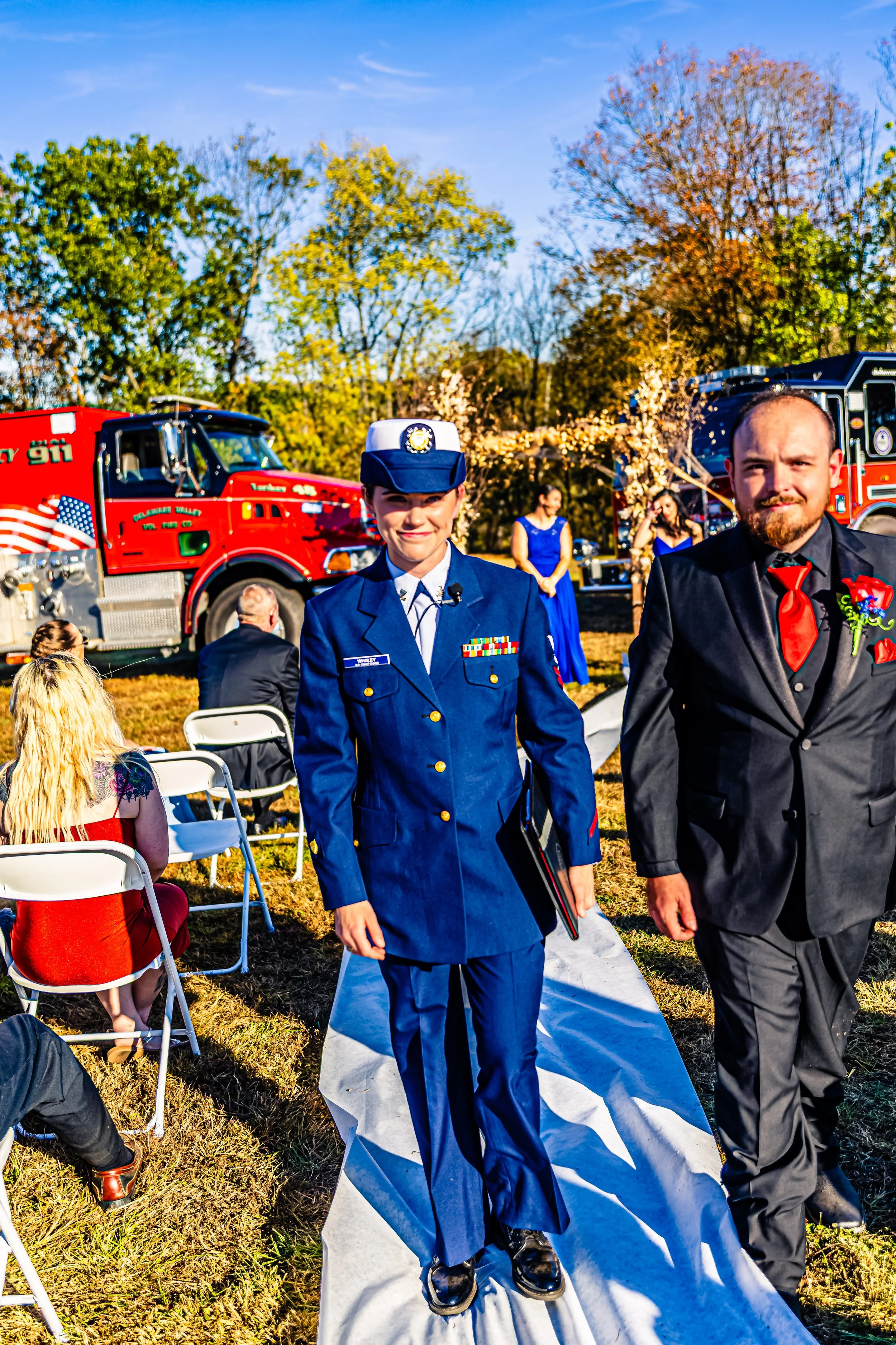 A woman in a military dress uniform and a man in a black suit with a red tie walking on a white aisle at an outdoor event, with a fire truck in the background and guests seated nearby.