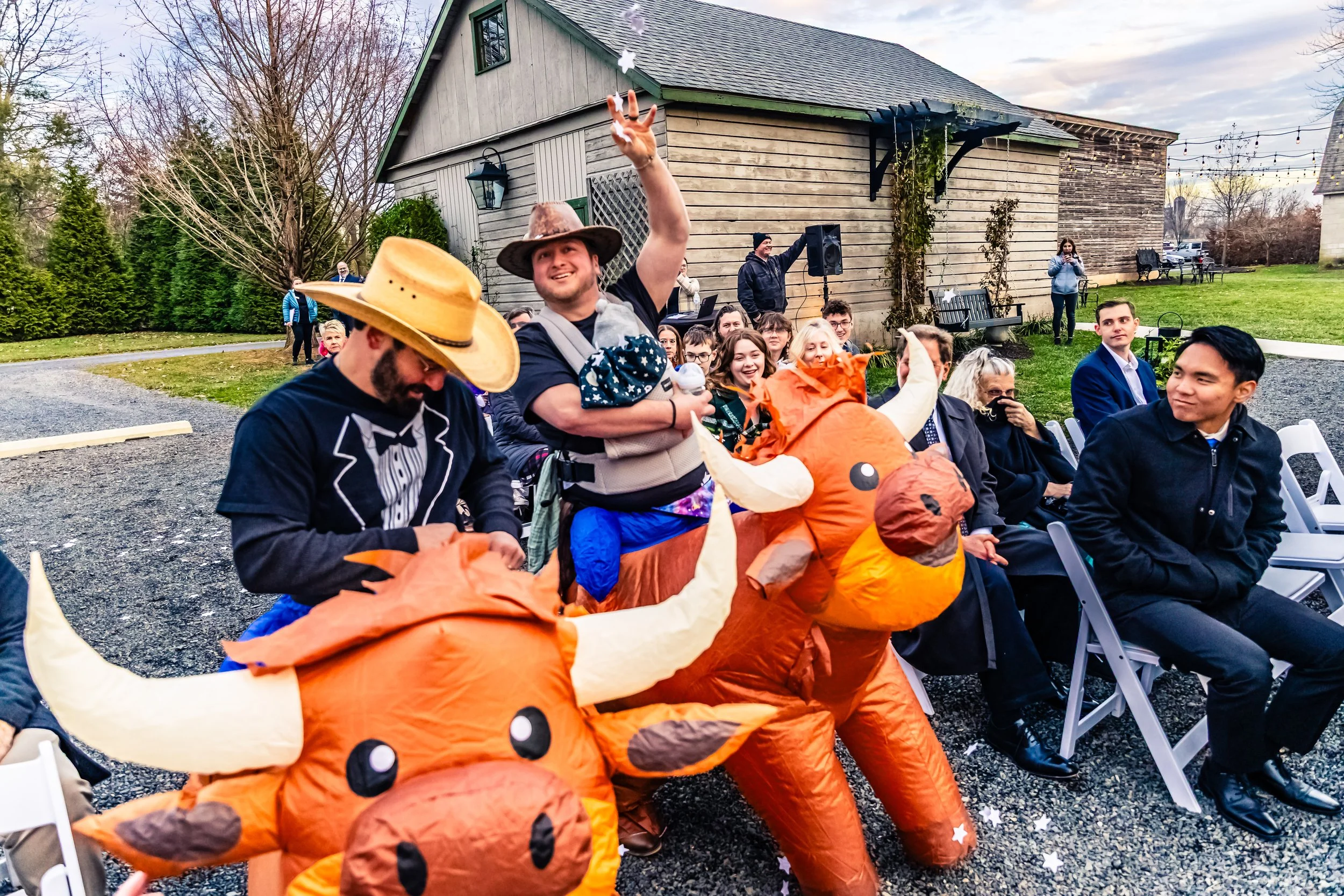 People attending outdoor celebration, with two individuals riding inflatable ox costumes, one person waving, and people sitting in chairs on a gravel ground near a wooden building with trees and string lights in the background.