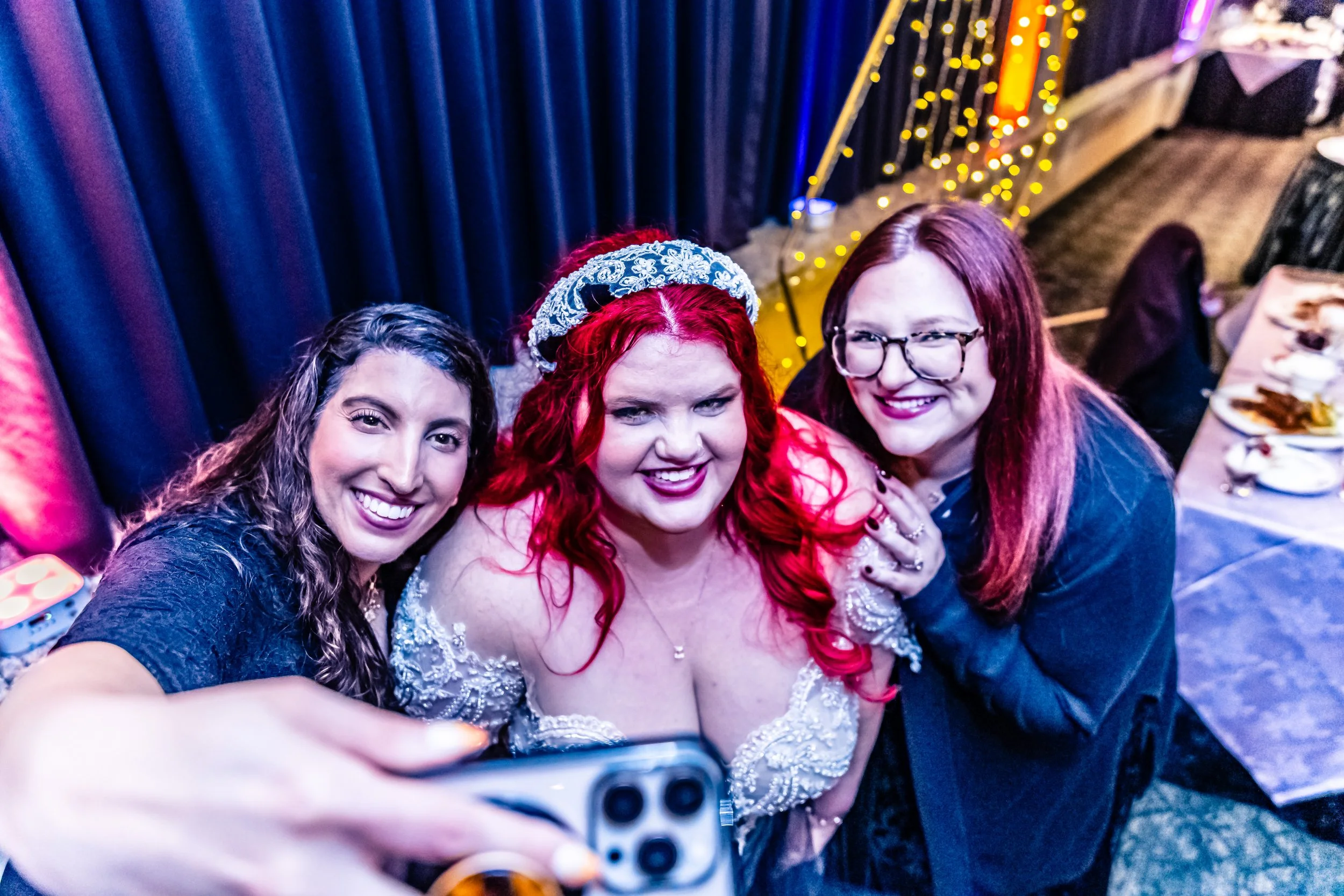Three women taking a selfie at a celebration event, with one woman dressed in a white dress and wearing a tiara, curly red hair, and a smiling expression, while the other two women, one with glasses and one with long brown hair, smile and pose with h