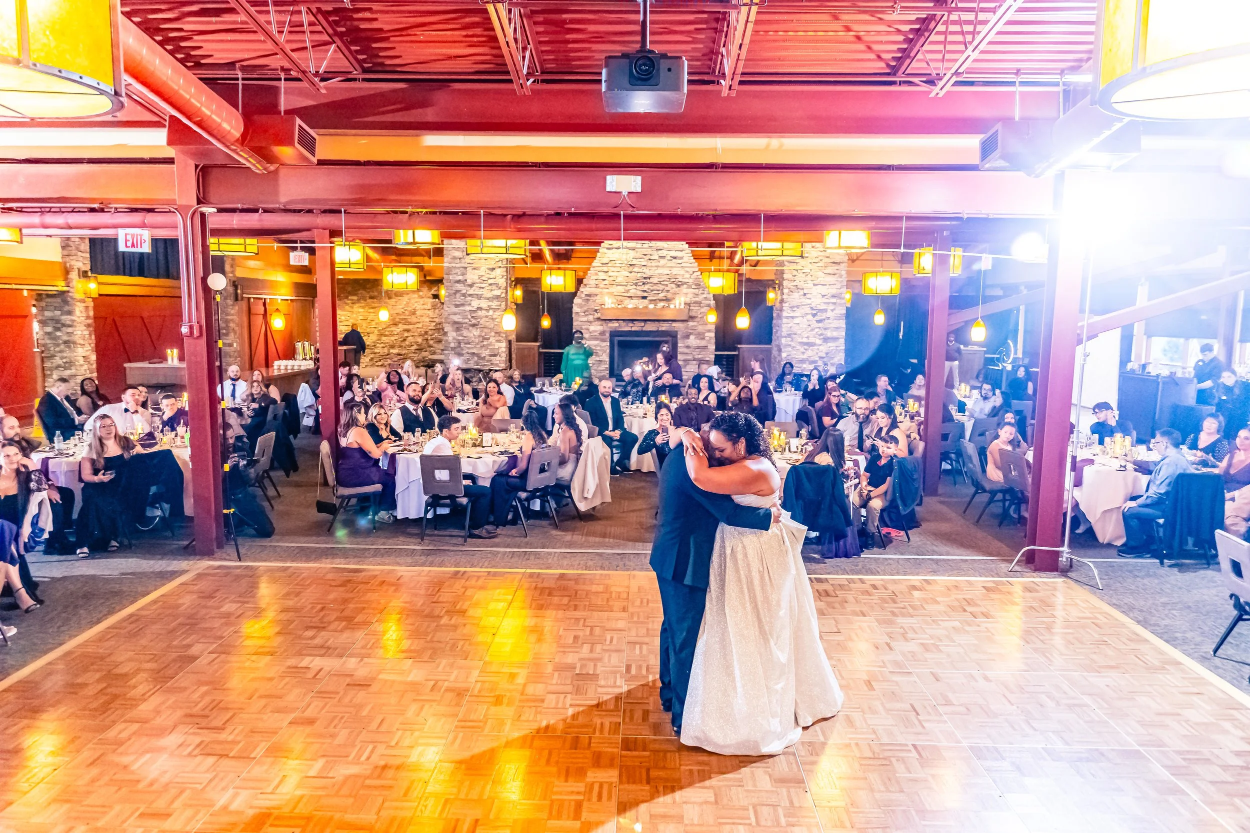A bride and groom dancing on a wooden dance floor at their wedding reception, with guests seated at round tables watching and celebrating in a warmly lit banquet hall.