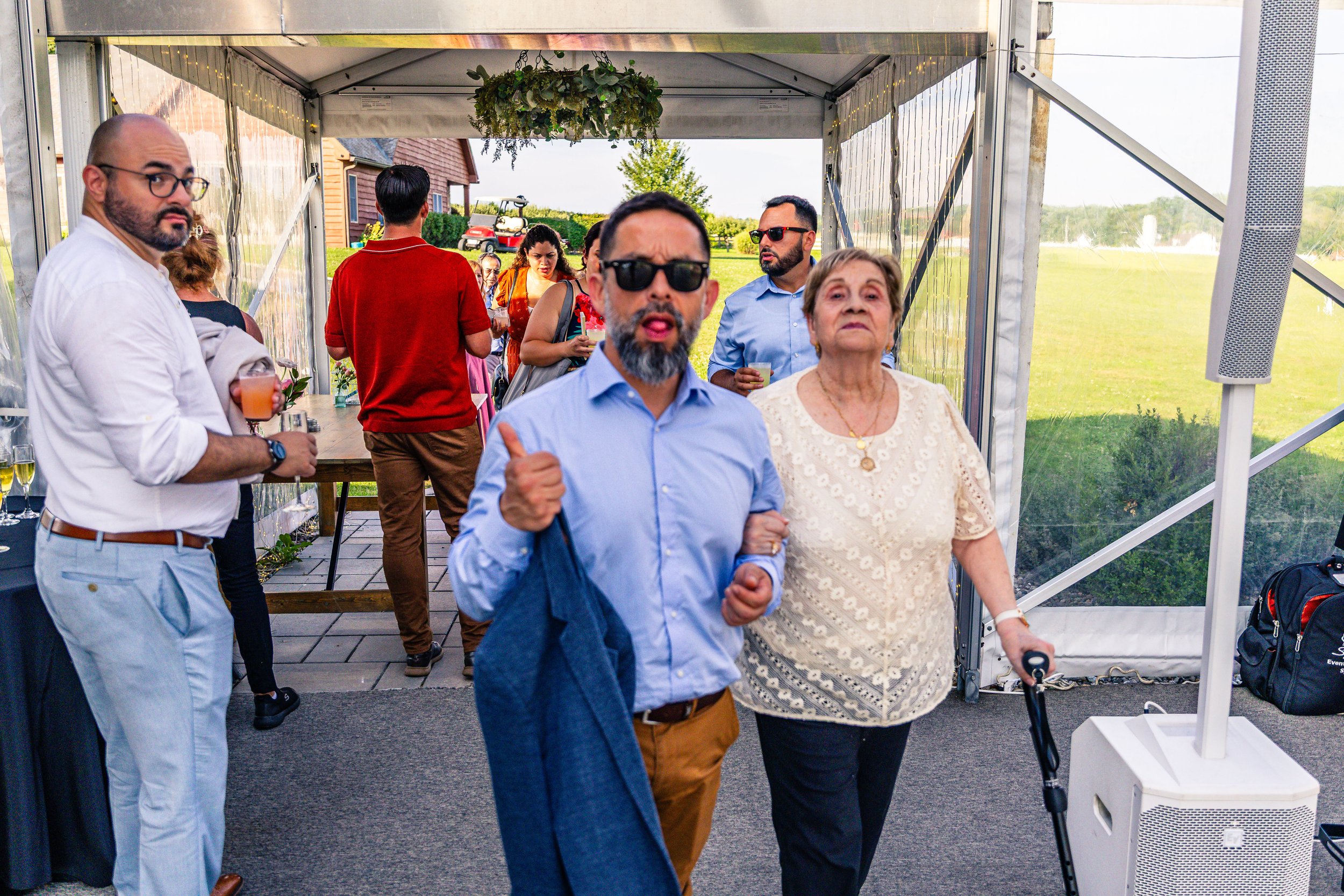 People attending an outdoor event inside a tent, with some holding drinks, and a man giving a thumbs-up while making an expressive face in the foreground.
