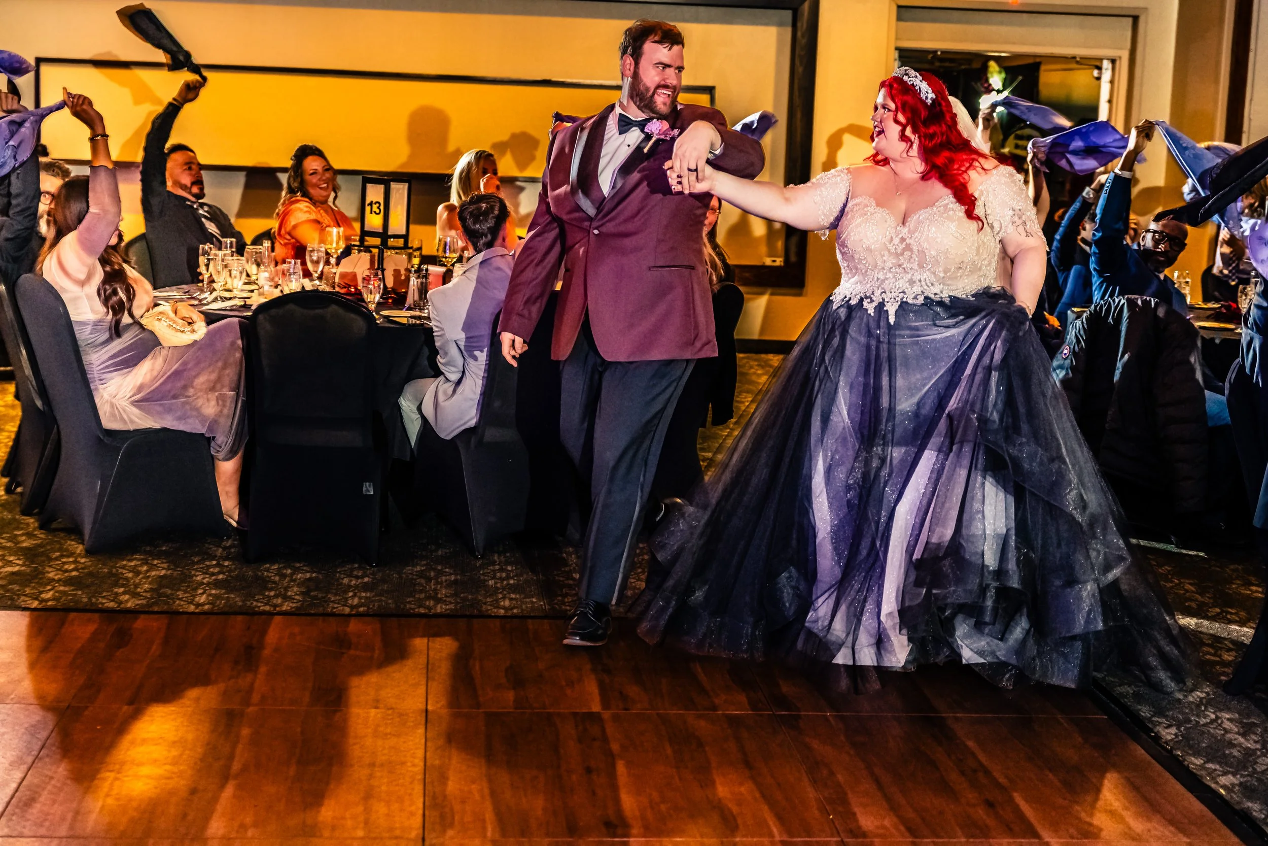A man and woman dance together at a wedding reception, with guests seated at tables watching and cheering in the background.