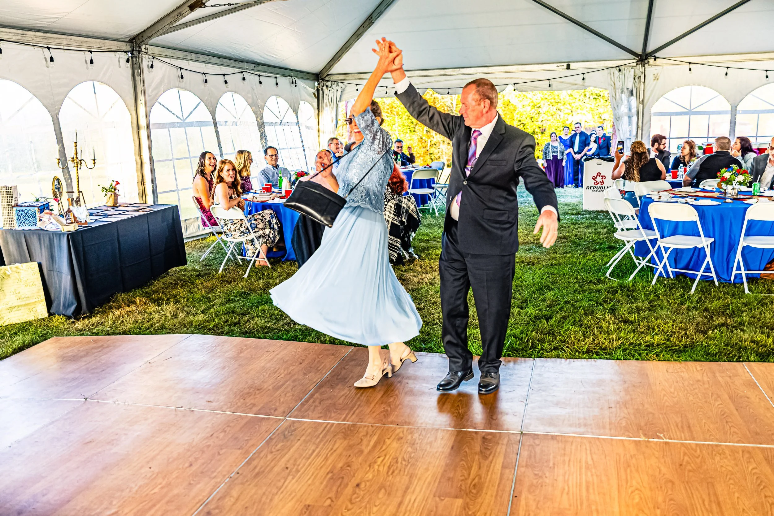 A man and woman dancing together at a wedding reception inside a decorated tent, with guests seated at tables watching.