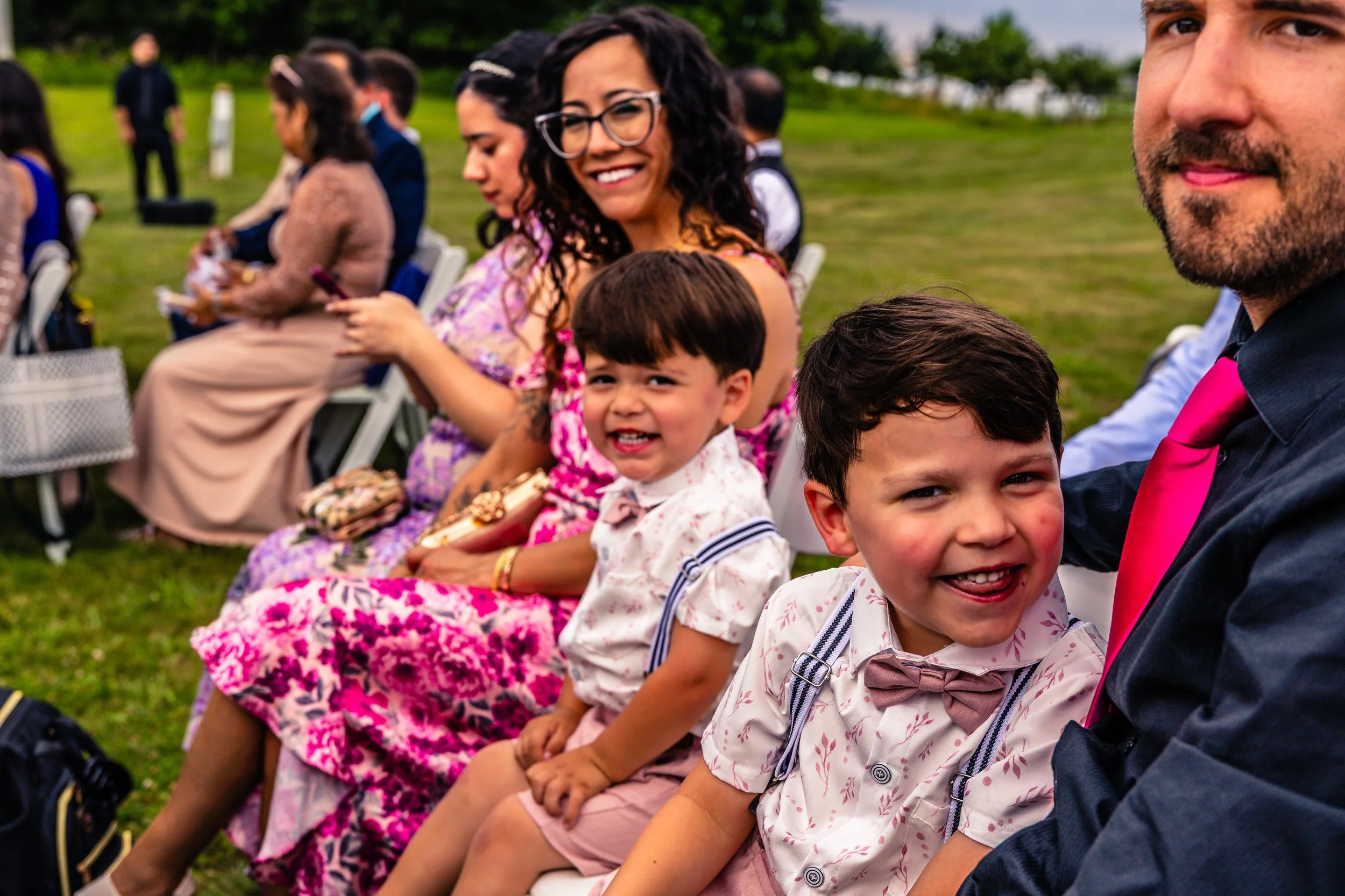A group of people, including children and adults, sitting outdoors on white chairs during daytime. The children in the foreground are smiling and sitting next to a man wearing a black shirt and pink tie. The background shows more people seated on the