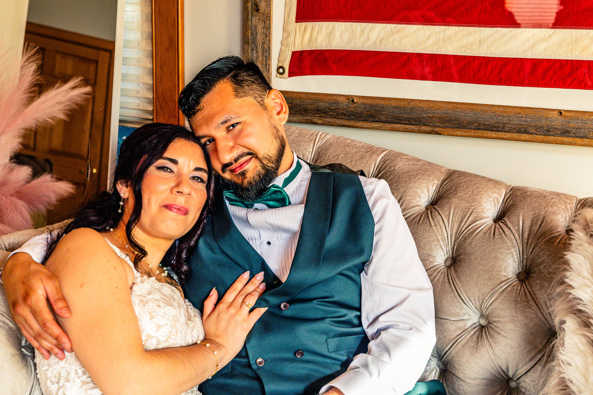 A couple sitting on a beige tufted sofa, smiling and embracing, with patriotic decor and a flag in the background.