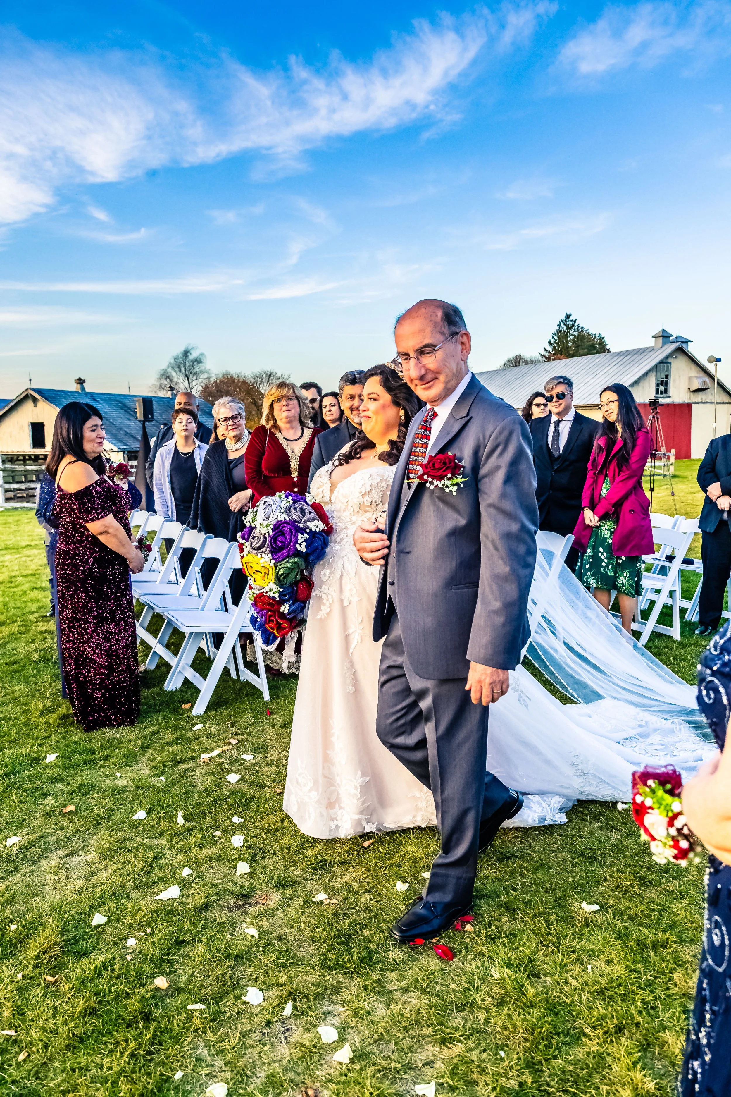 A bride and an older man, likely her father, walking down the aisle at an outdoor wedding ceremony, with guests seated on either side on a grassy field under a partly cloudy sky.