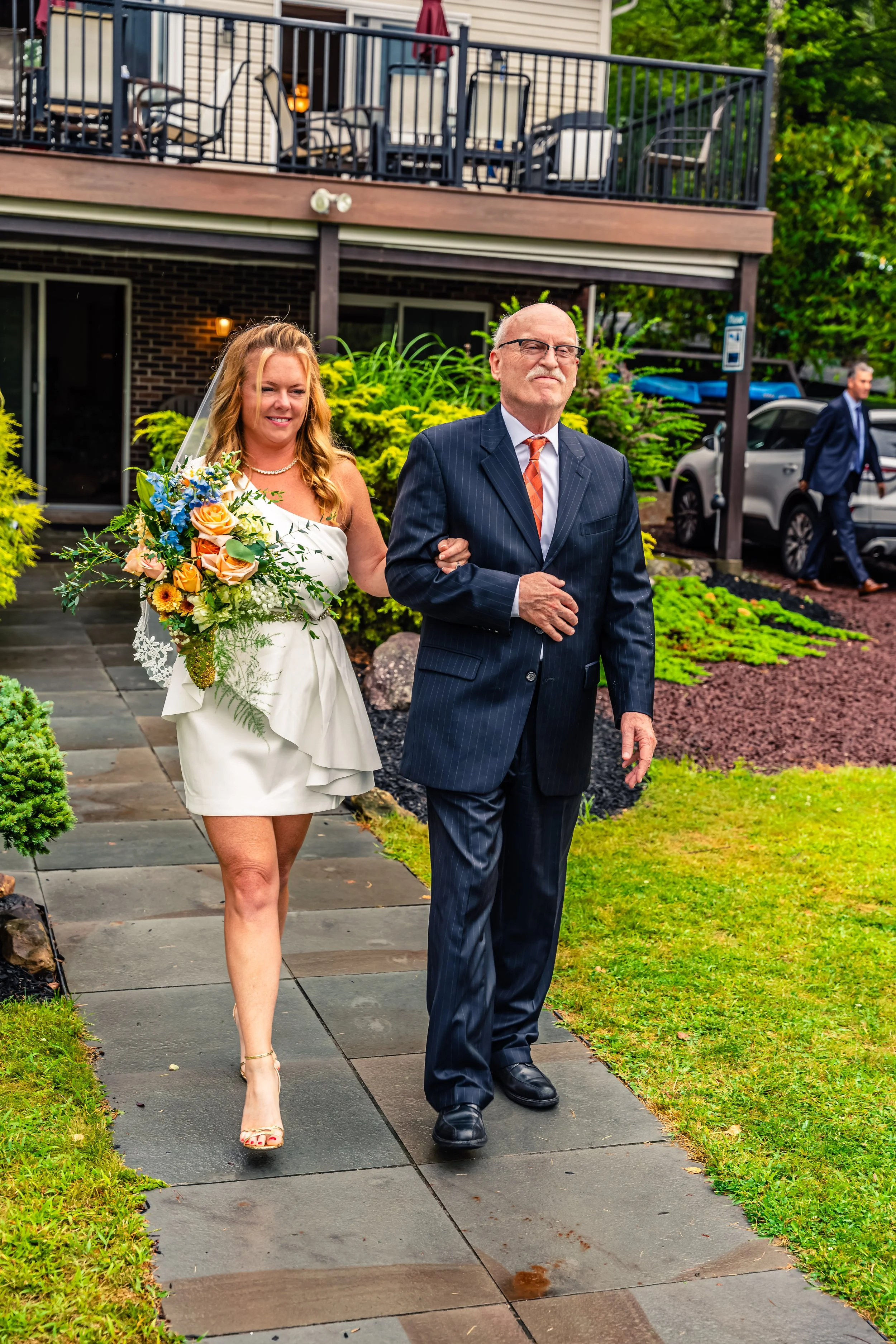 A bride in a white dress holding a colorful bouquet, walking arm-in-arm with an older man in a dark suit outside a building with a patio and parking area.