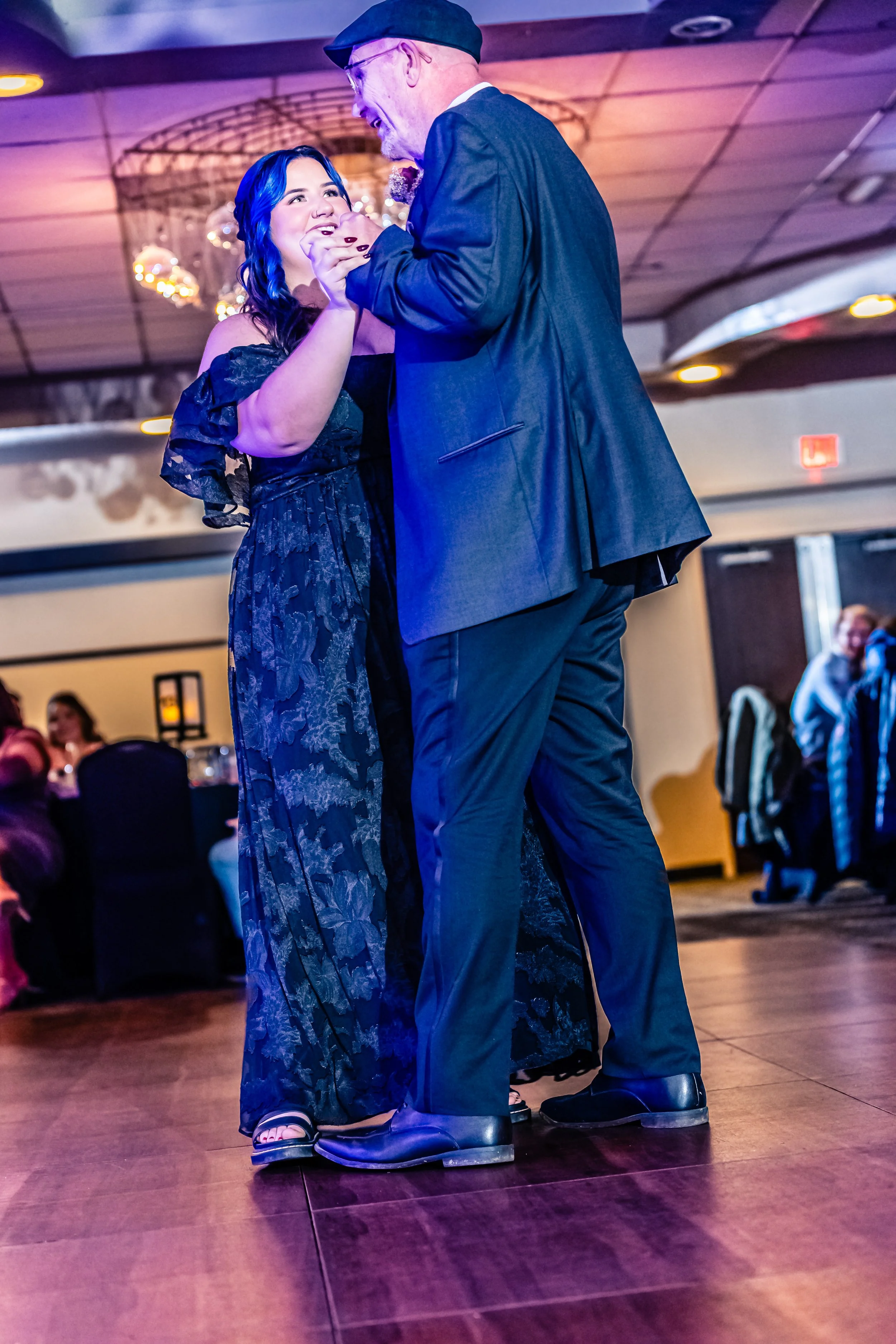 A woman with blue hair and a black dress dancing with an older man in a suit and cap at a social event or dance floor, with other guests in the background.