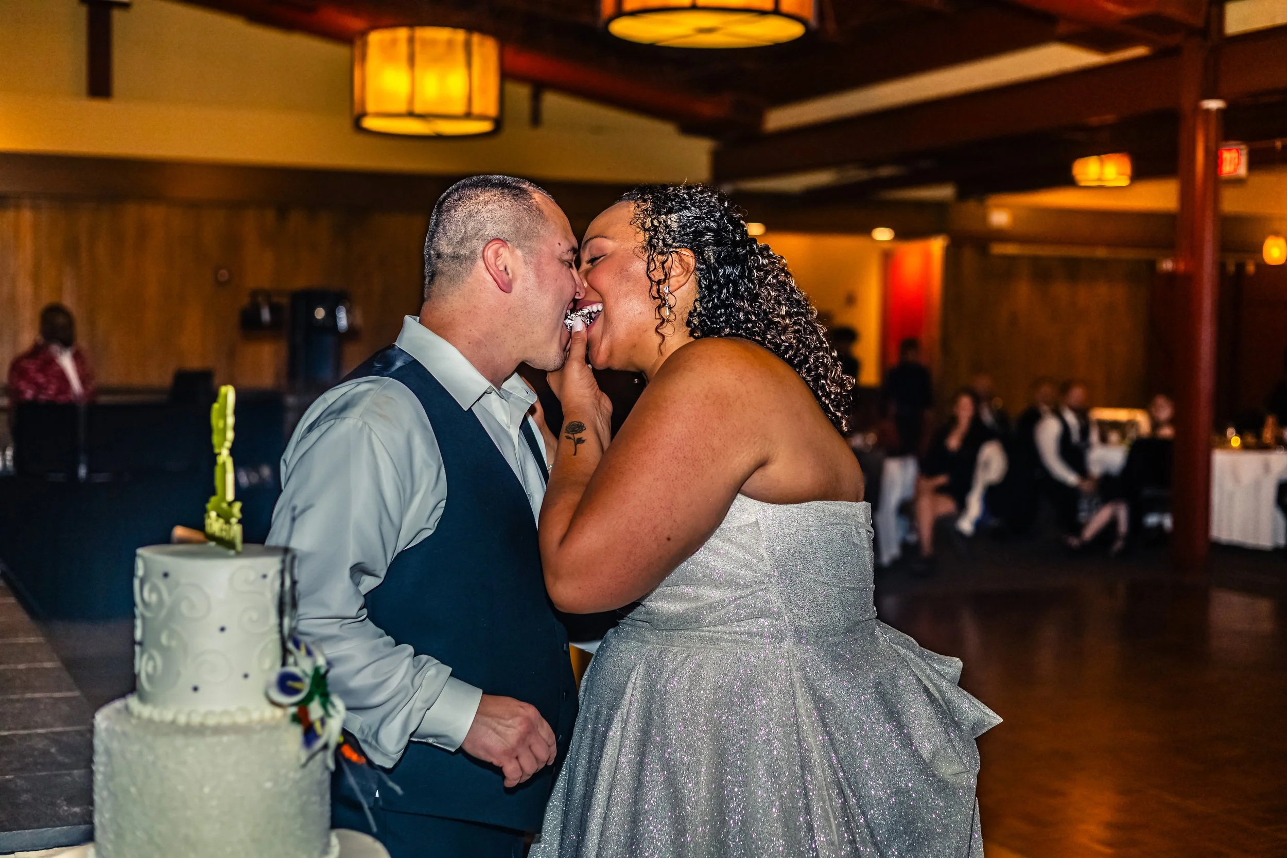 A couple is sharing a kiss at their wedding reception, with a wedding cake visible in the foreground and guests seated at tables in the background.
