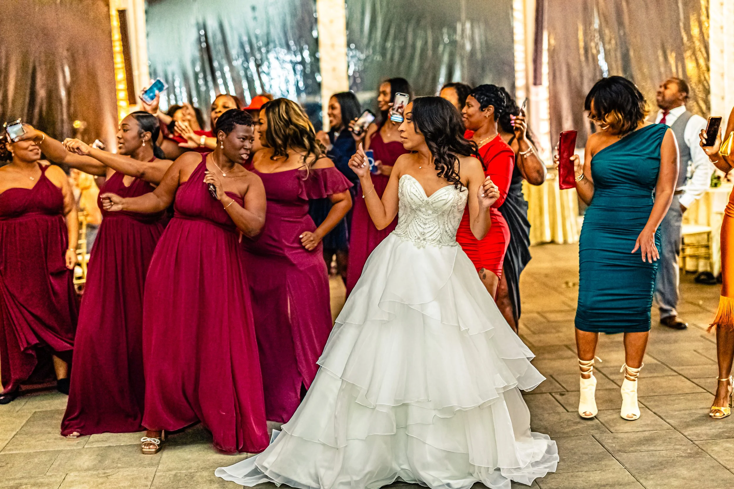 Wedding reception with a bride in a white gown dancing among women wearing maroon, red, teal, and other colorful dresses, some holding drinks and smartphones.