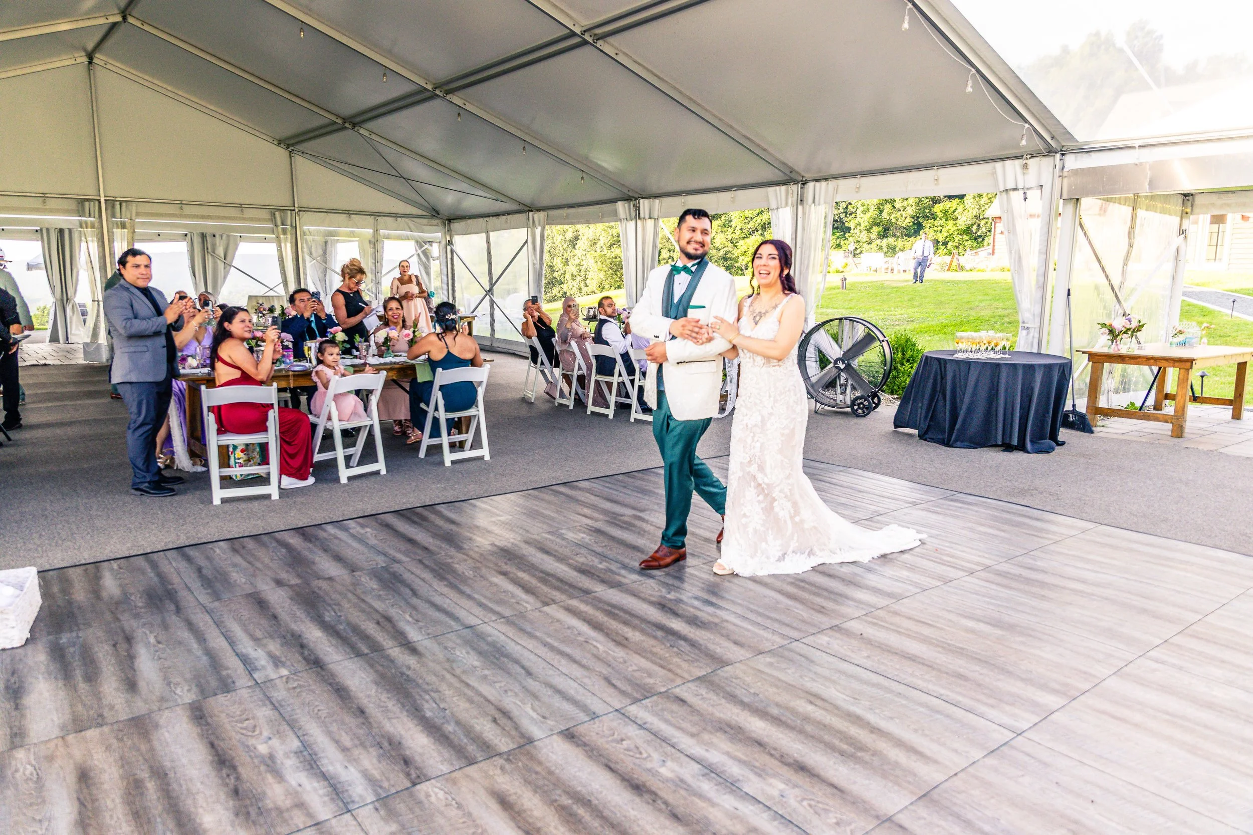 A bride and groom dancing at their wedding reception inside a large tent with a wood floor. Guests are seated at tables, some taking photos, and outdoor greenery is visible in the background.
