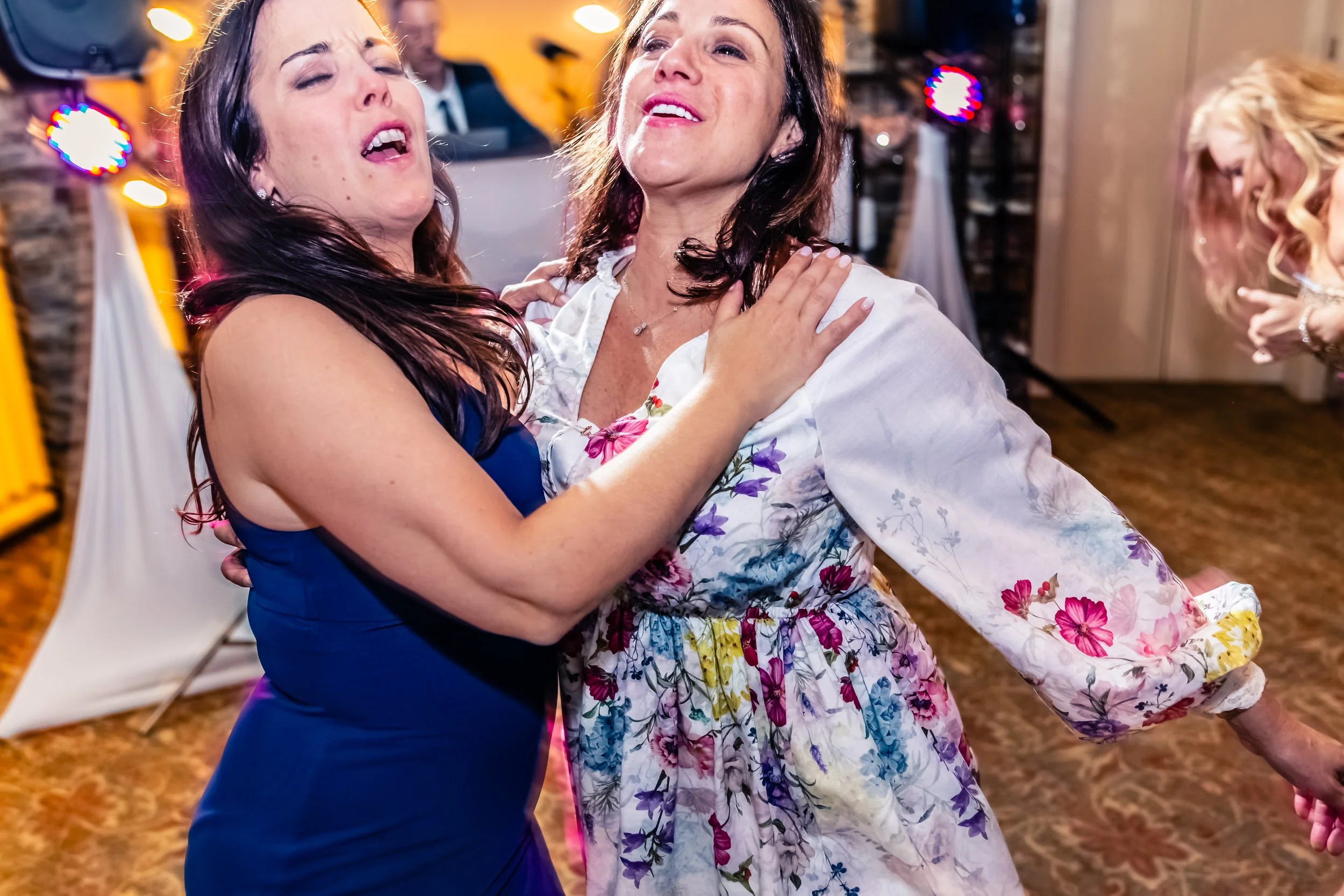 Two women dancing closely together at a celebration or party, with a blurred DJ and colorful lights in the background.