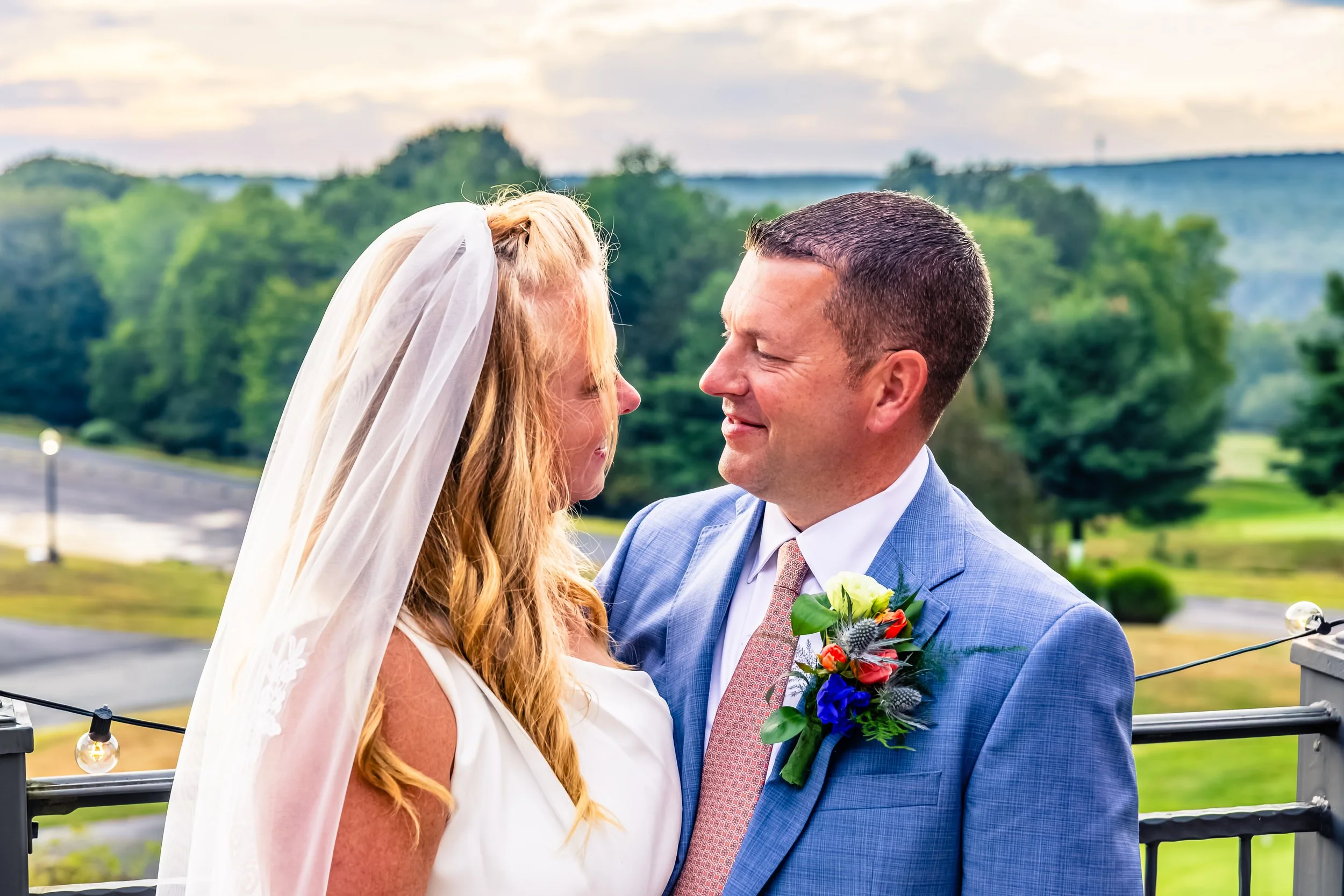 A bride and groom stand close together, smiling and gazing into each other's eyes outdoors during their wedding ceremony, with green trees and a cloudy sky in the background.