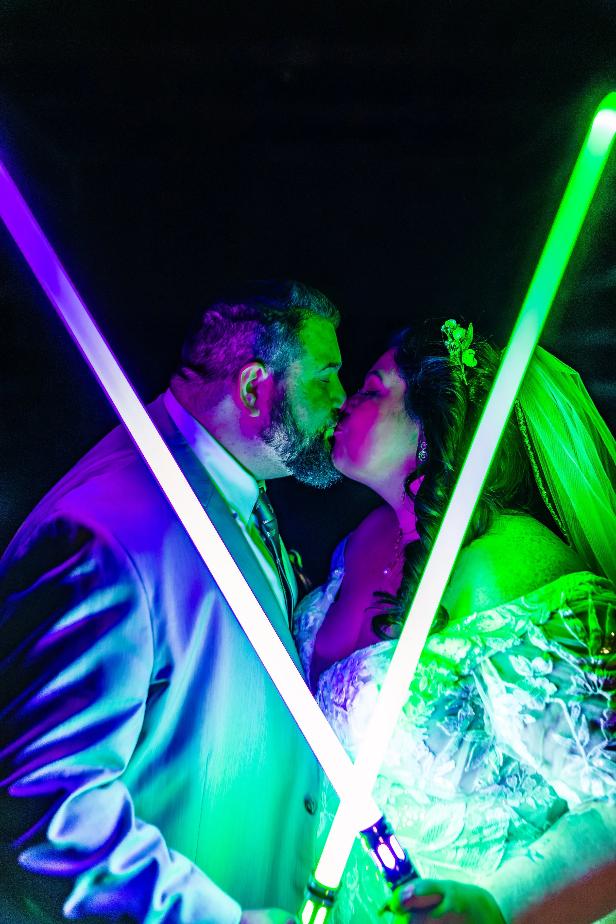 A newlywed couple, dressed in wedding attire, kiss under colorful neon lights, with the groom in a gray suit and the bride in a white gown with a veil.