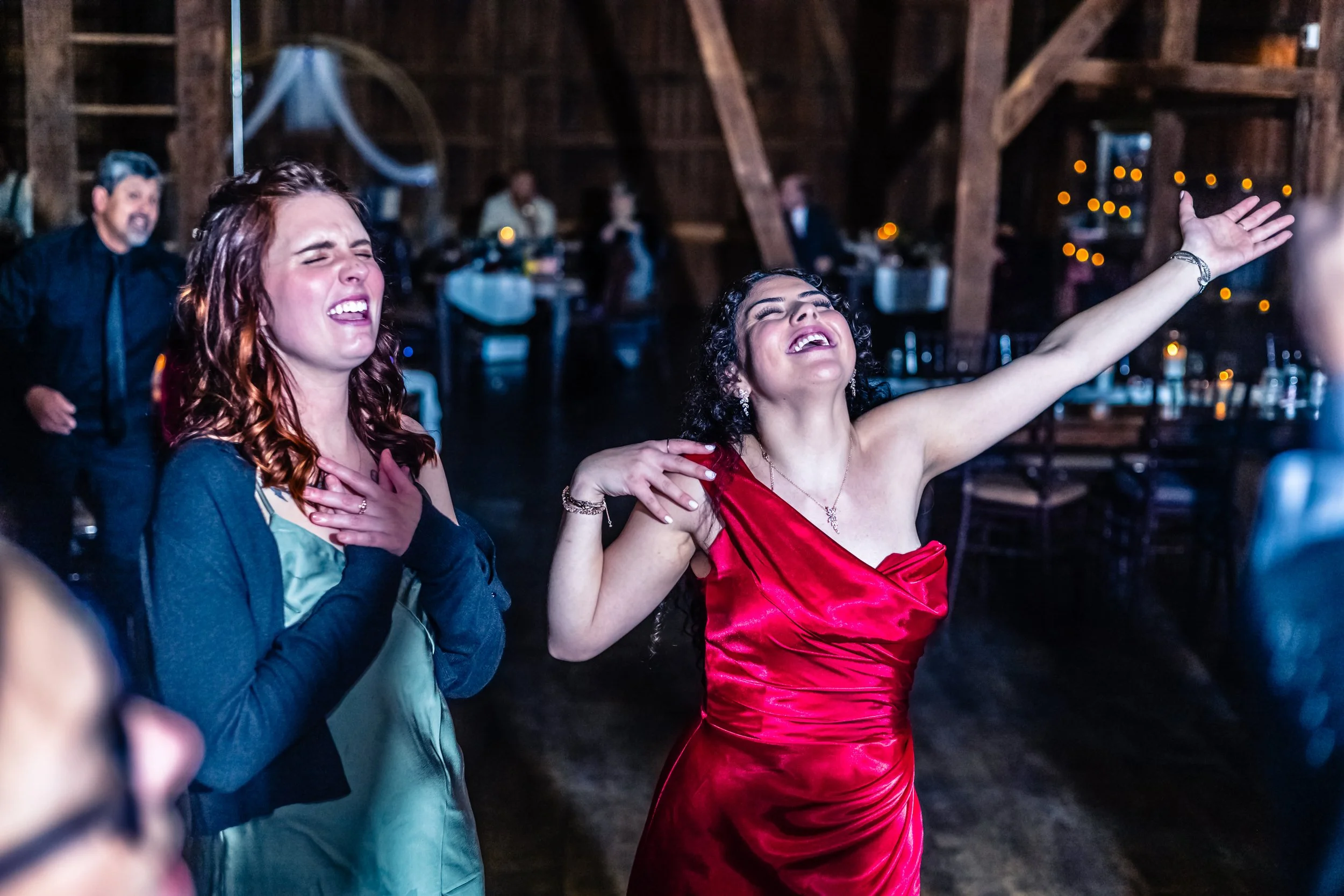 Women dancing at a celebration or wedding reception inside a rustic wooden venue, with joyful expressions and stylish attire.