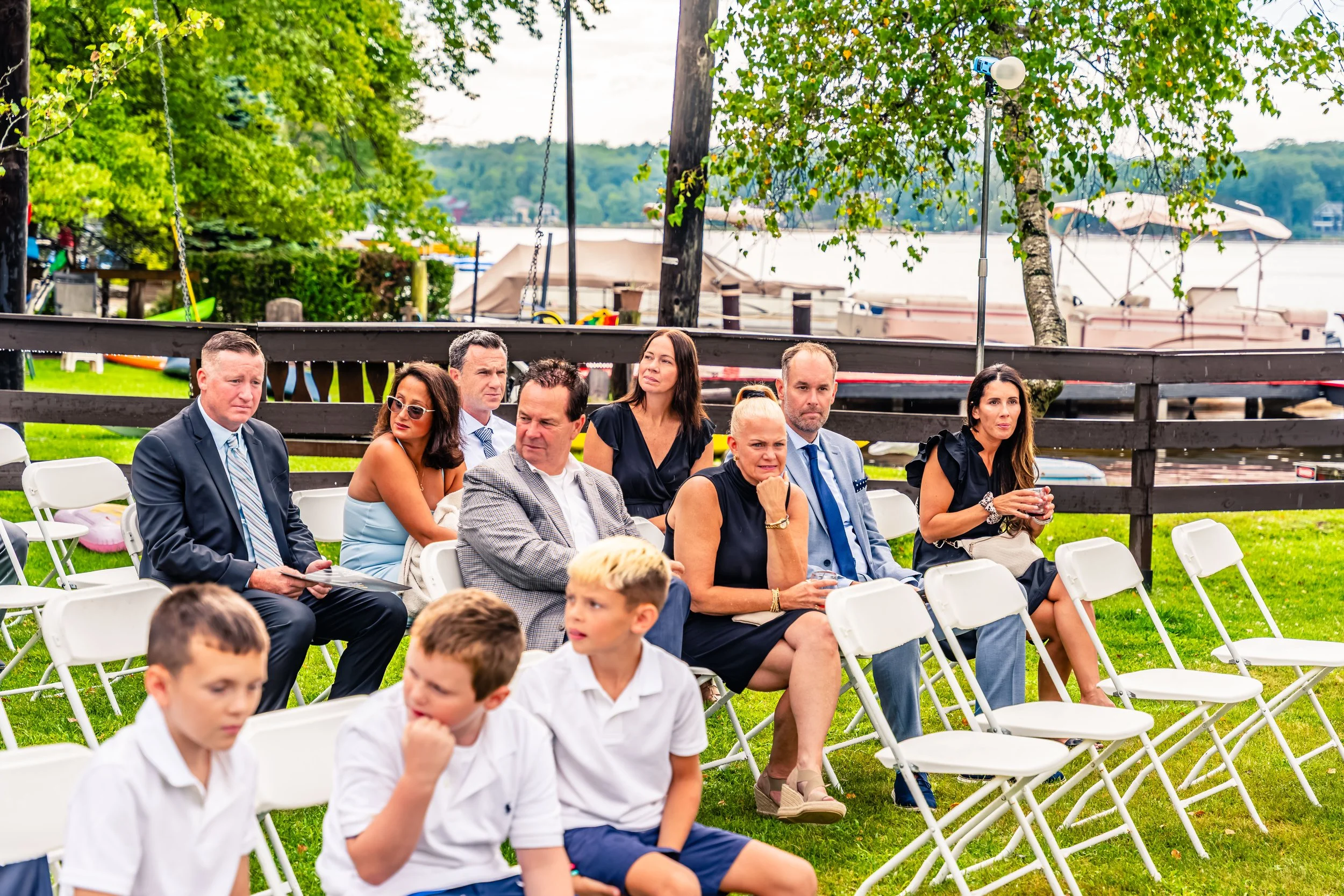 People sitting on white chairs outdoors near a lake, attending an event, with trees and boats in the background.