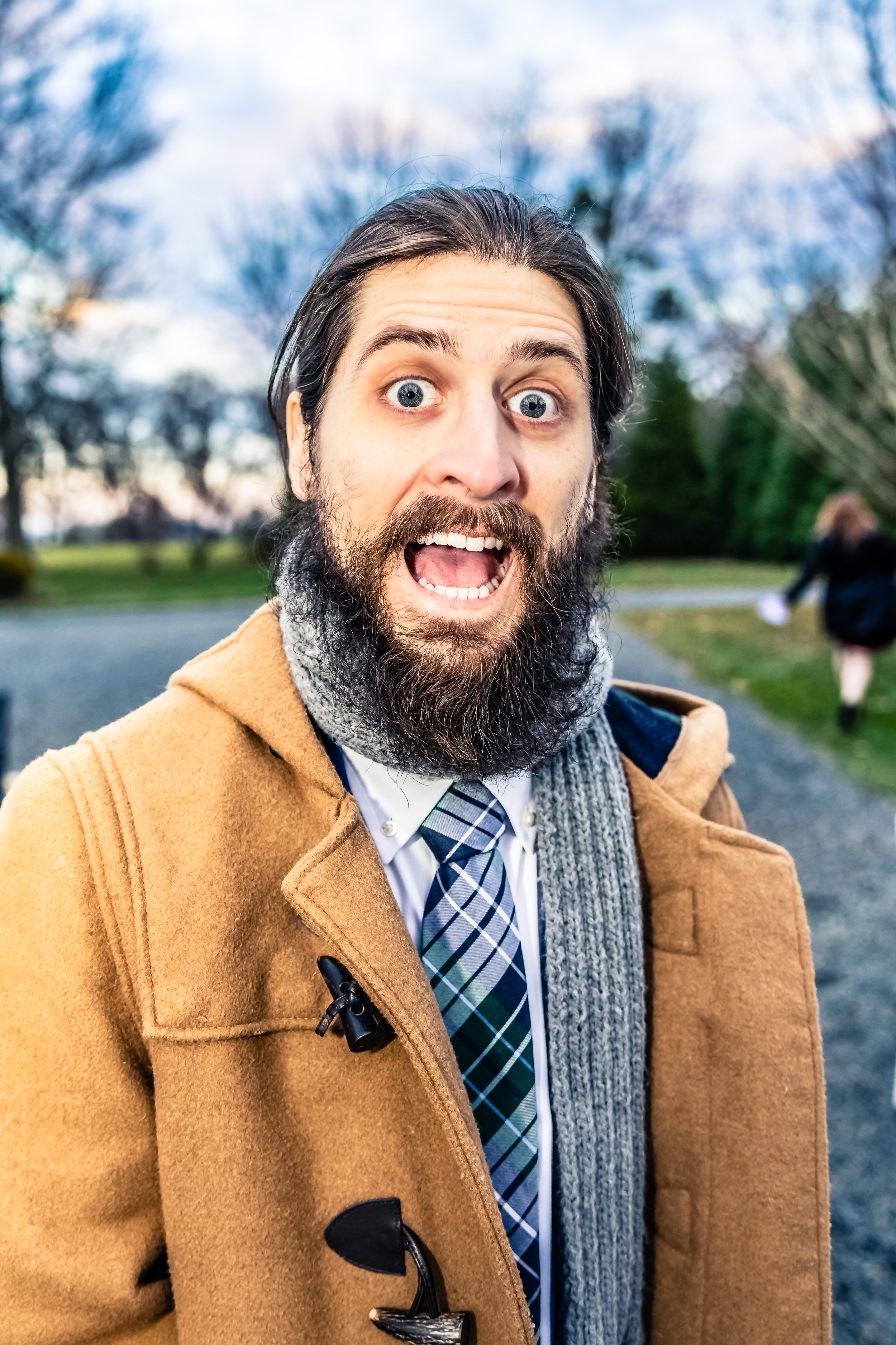 A man with a beard and long hair wears a brown coat, a gray scarf, a white shirt, and a plaid tie, standing outdoors with trees and a person walking in the background, making a surprised facial expression.