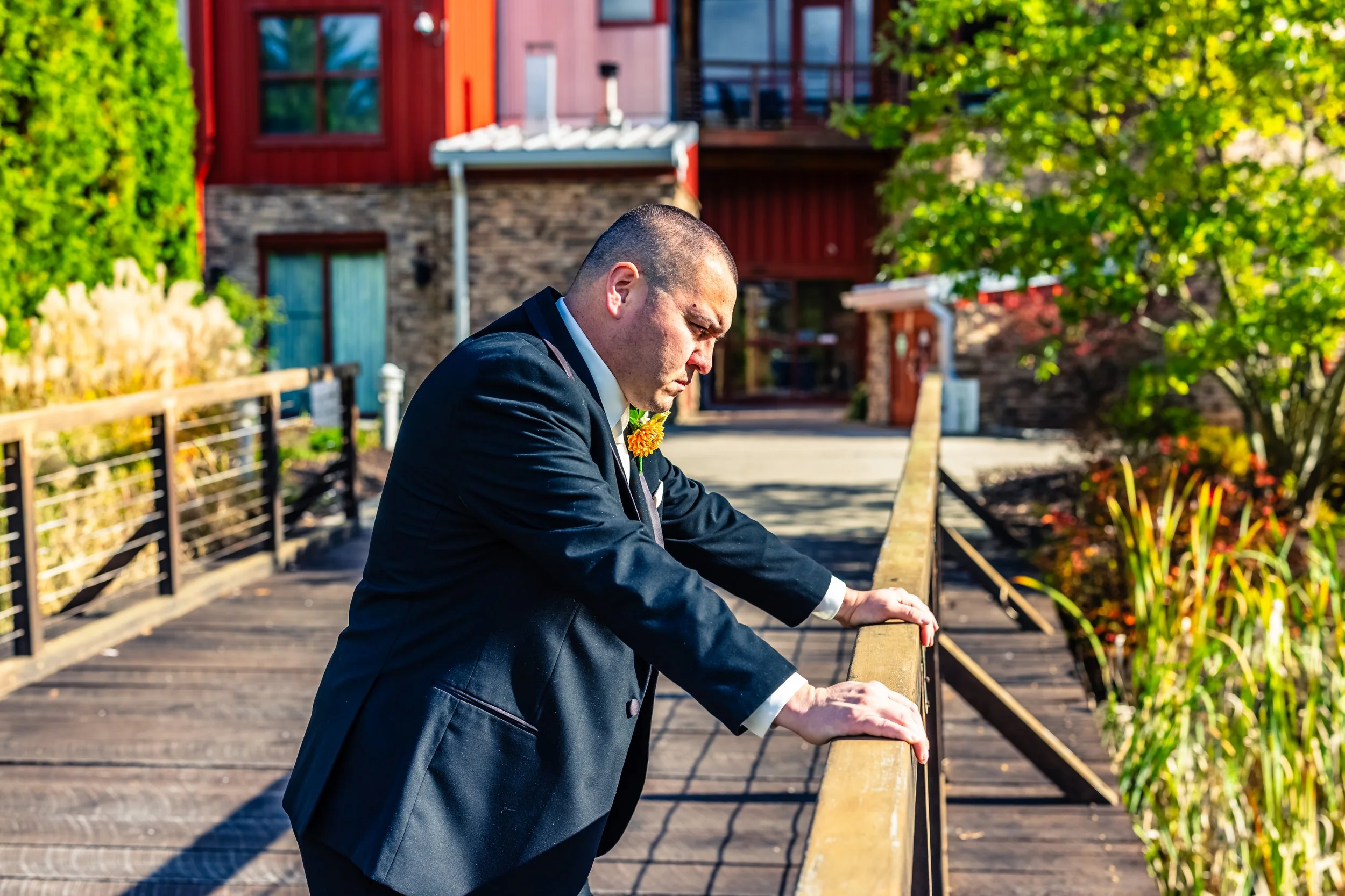A man in a dark suit leans on a wooden railing on a bridge, looking down thoughtfully, with a red building and greenery in the background on a bright sunny day.