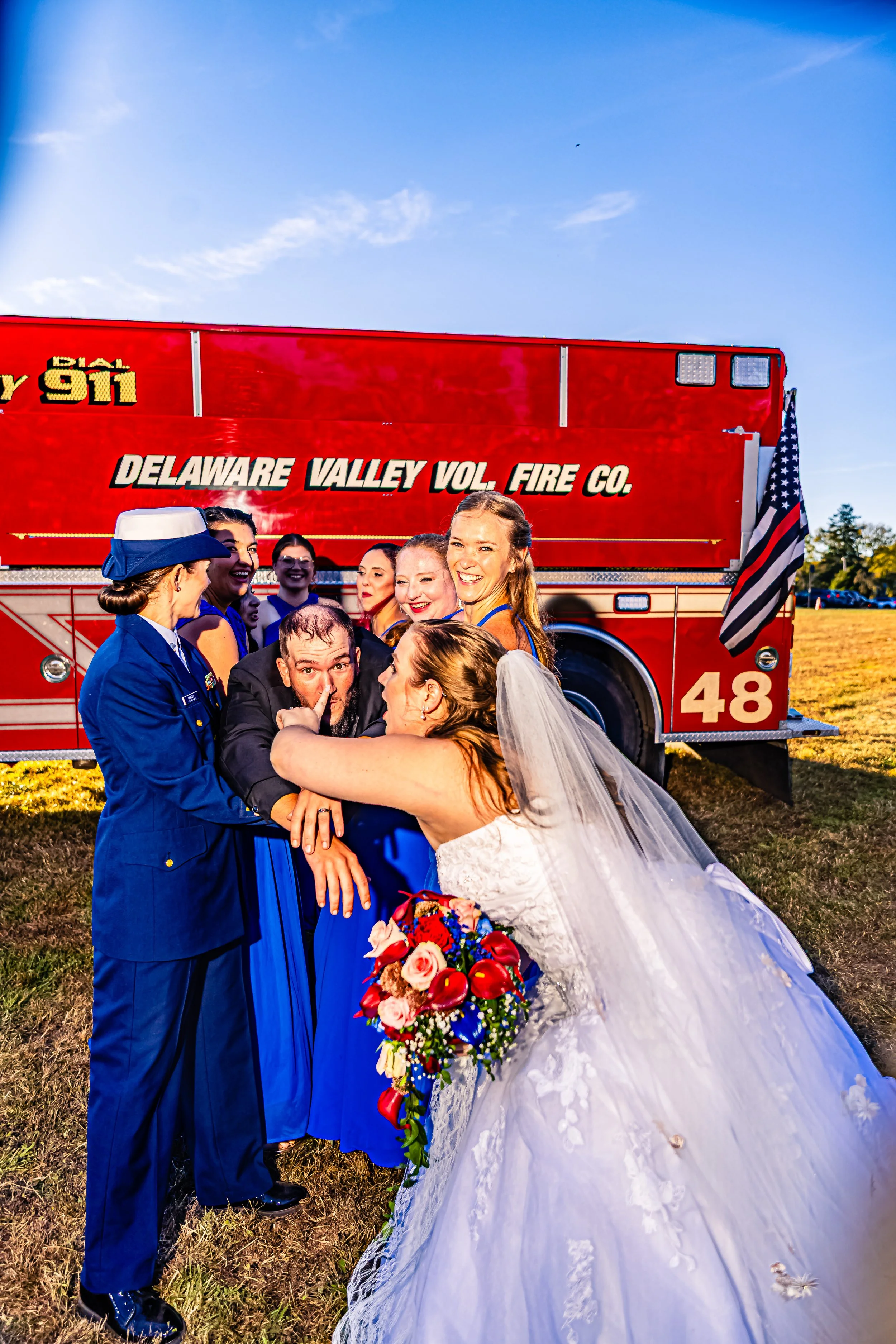 A bride in a wedding dress kissing a firefighter, surrounded by friends and a firefighter in uniform, in front of a red fire truck with the text 'Delaware Valley Vol. Fire Co.' on it.