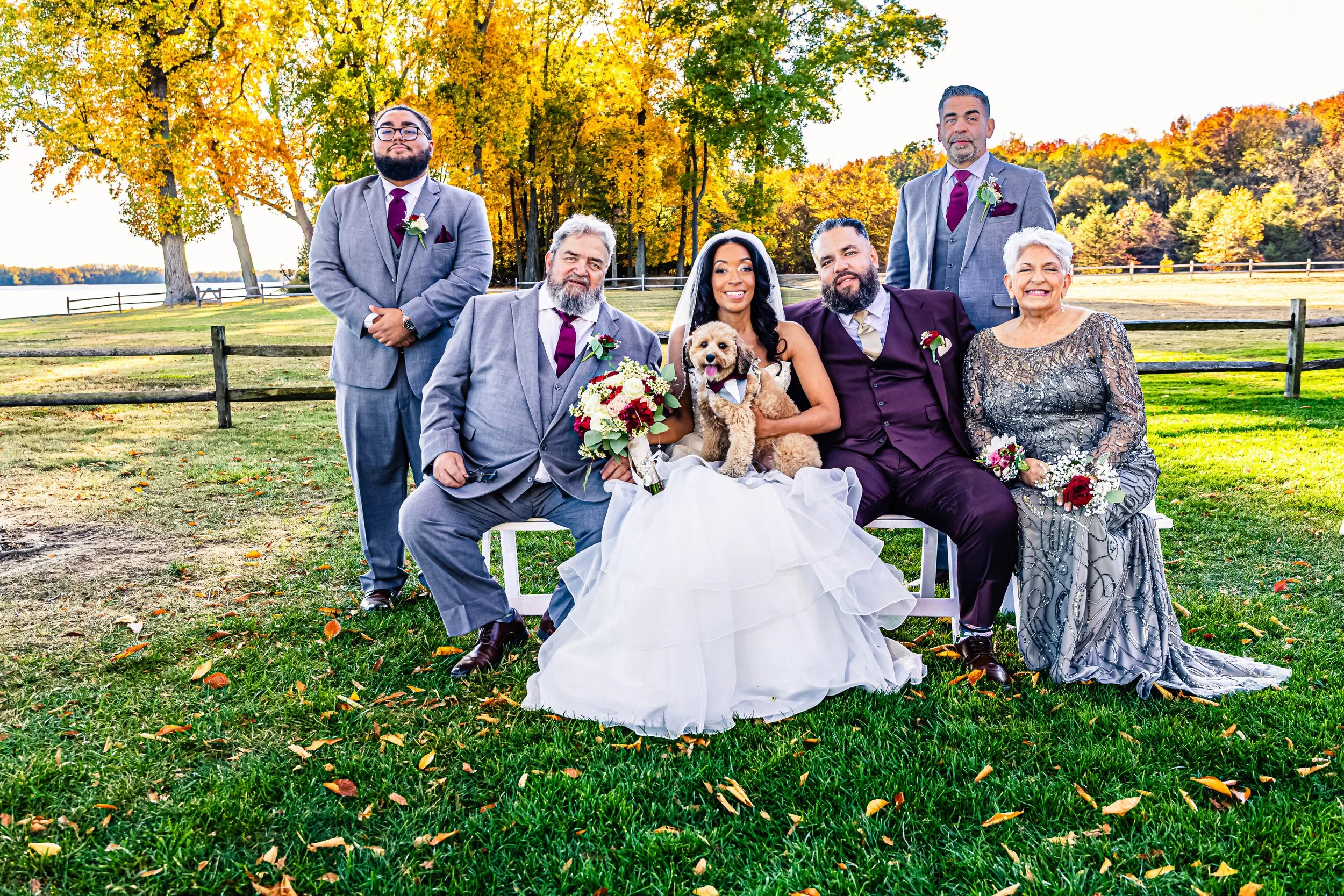 A group of six people and a dog at a wedding outdoors during autumn. The bride is sitting in the center, holding a bouquet and a small, fluffy dog. The groom is sitting next to her, and the others are standing or sitting nearby, all dressed in formal
