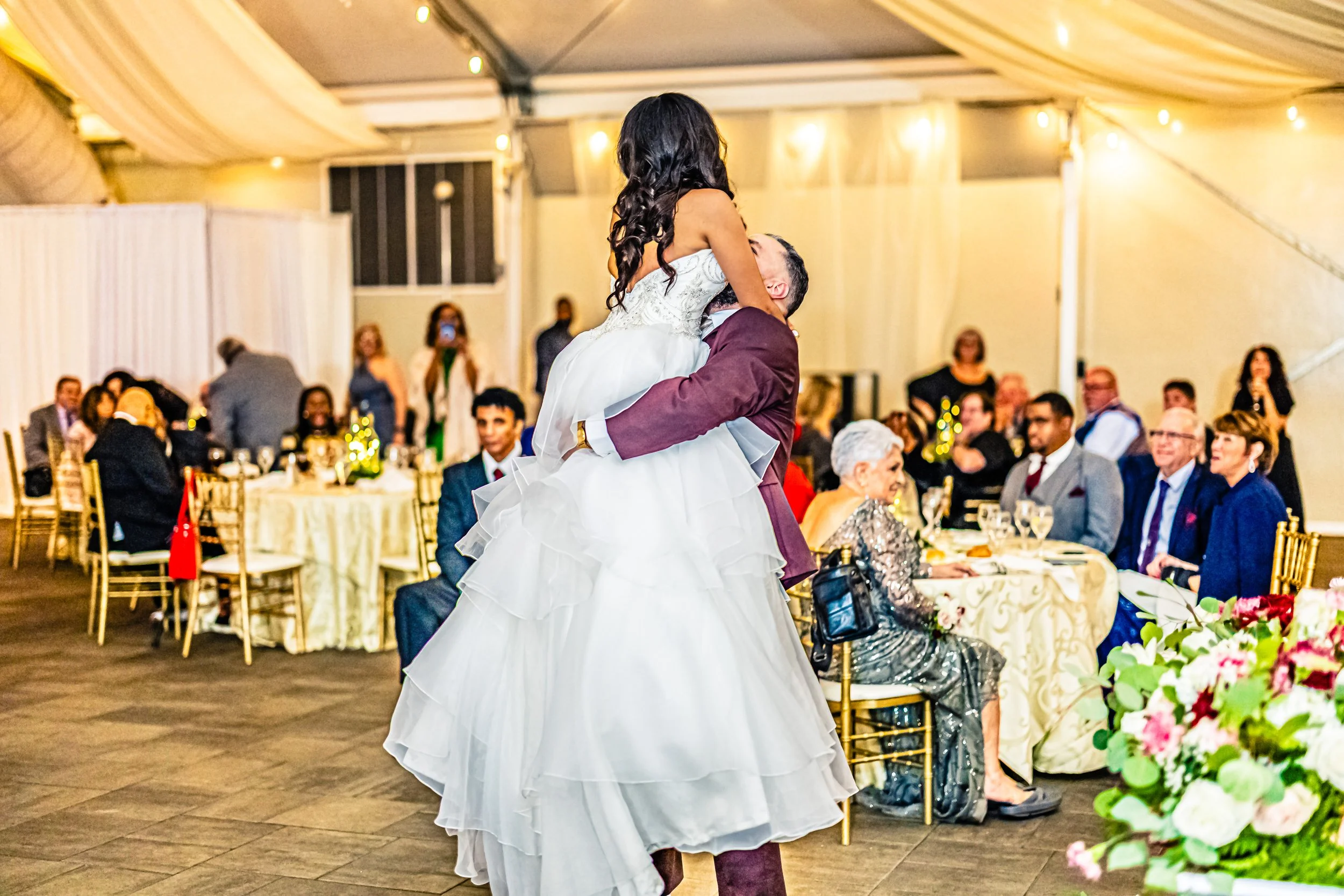 A bride and groom dancing at their wedding reception, the bride in a white gown lifted by the groom in a burgundy suit, guests seated at decorated tables in a large tent with warm lighting.