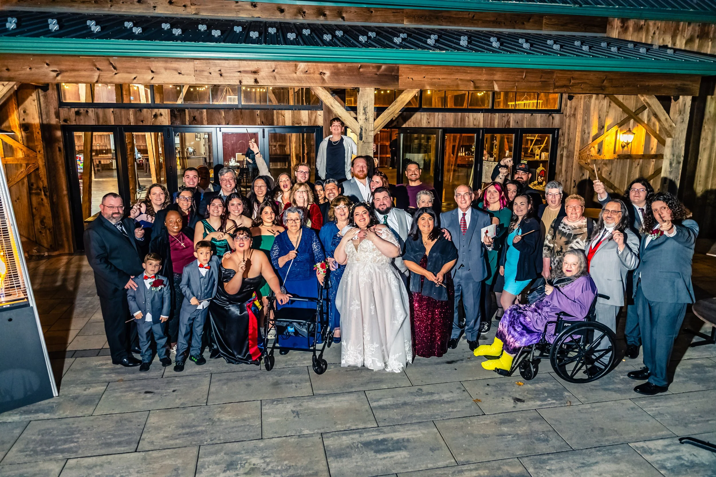 Group of people gathered outside of a rustic building at night, celebrating a wedding. The bride in a white wedding dress is in the center, surrounded by family and friends of various ages, some in formal attire and others in colorful outfits. Some i
