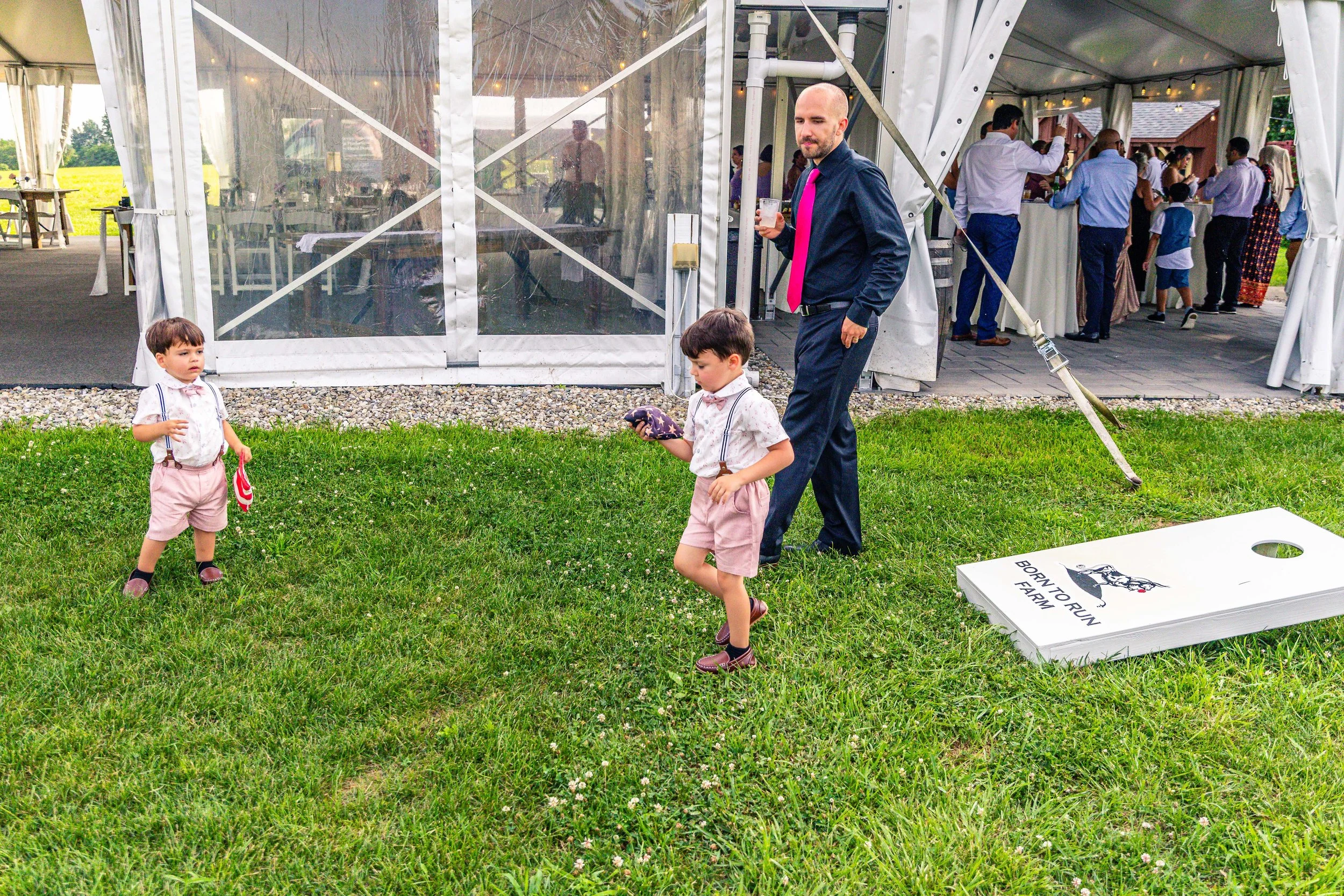 Kids playing cornhole game outside at a wedding reception, with a large tent and guests in the background.