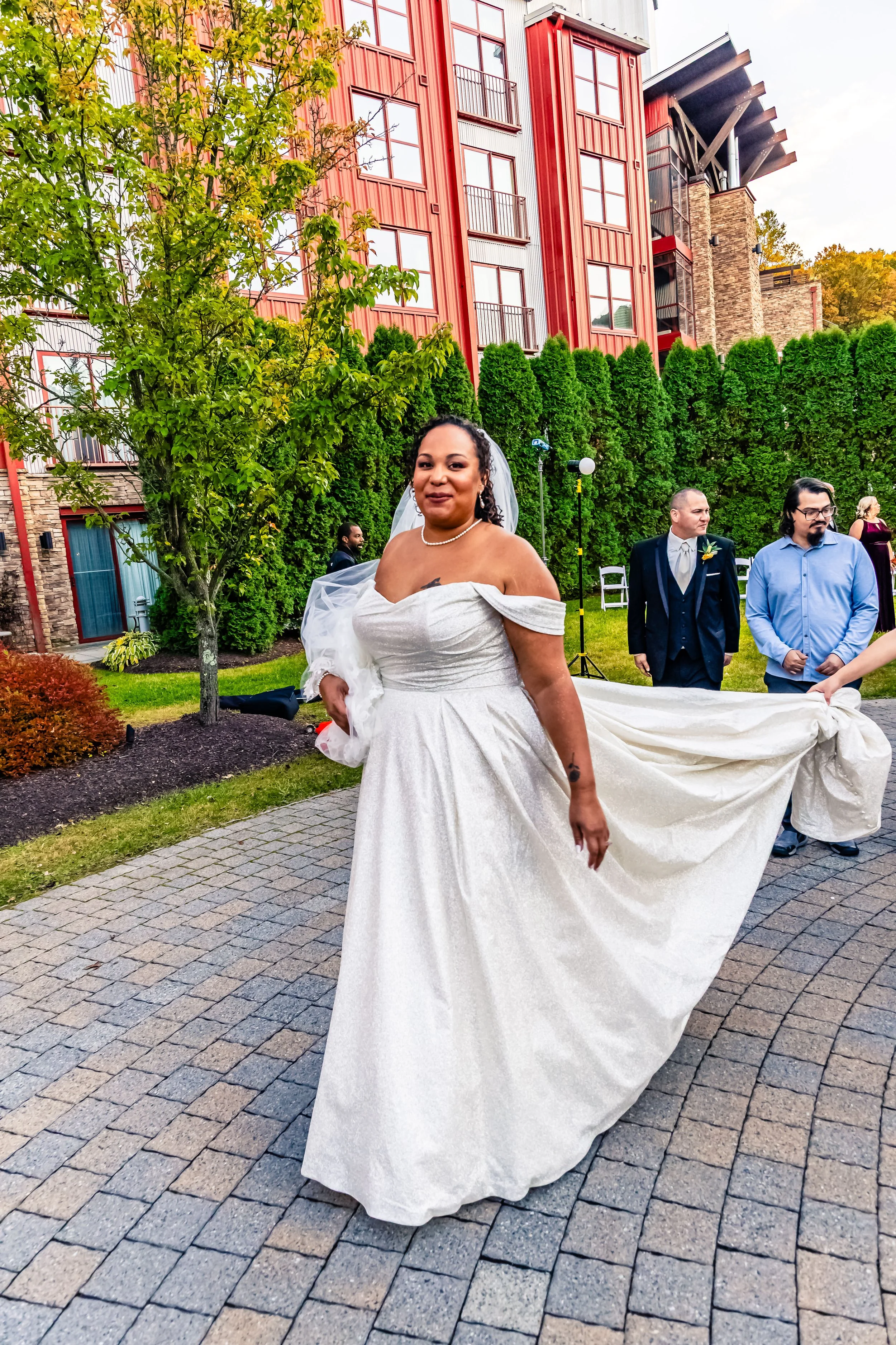 A bride in an off-shoulder white wedding gown standing outdoors on a paved pathway, with trees and a red building in the background.
