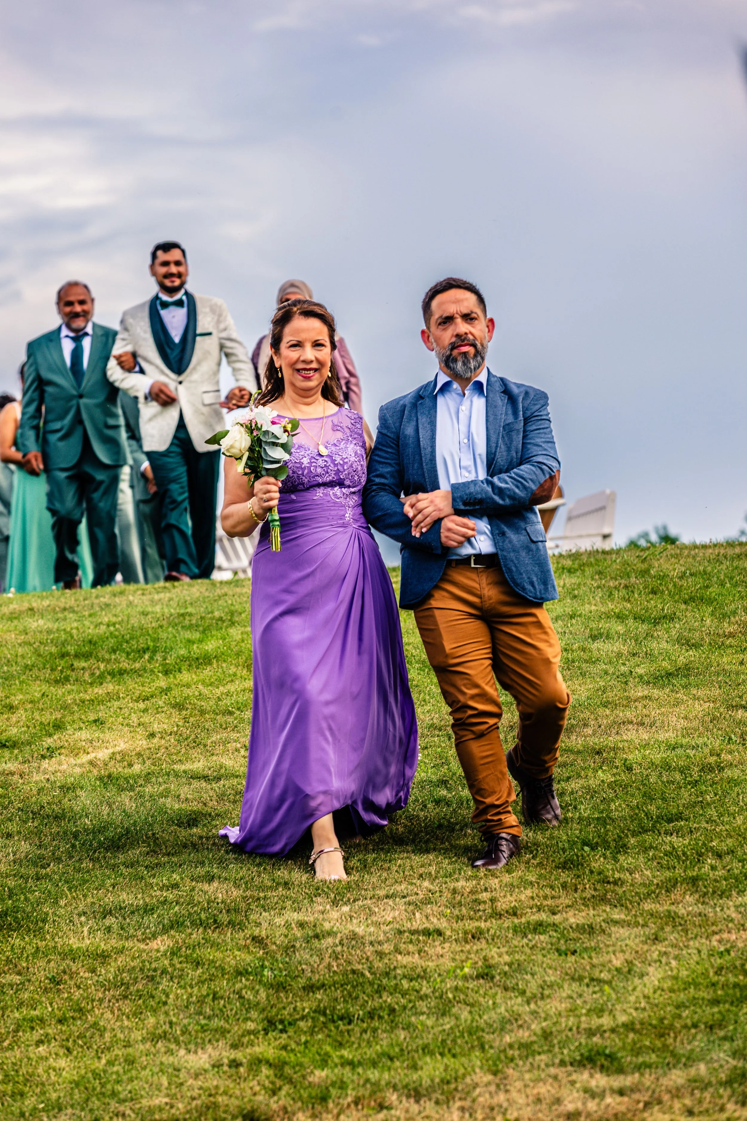 A group of people at a wedding, with a woman in a purple gown holding a bouquet and walking arm-in-arm with a man in a blue blazer and brown pants, outdoors on a grassy area under a cloudy sky.