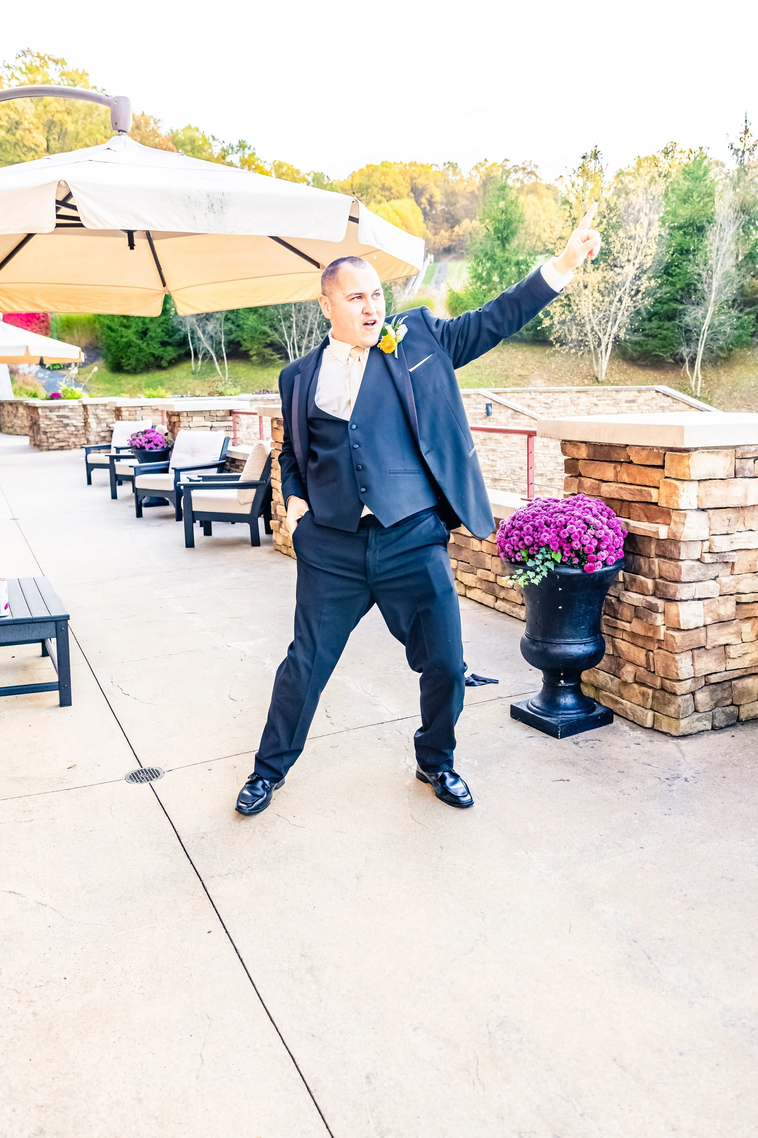 A man in a dark suit and tie, standing outdoors on a patio, gesturing upwards with his right hand, with a scenic background of trees and a stone wall, and large umbrellas covering seating areas.
