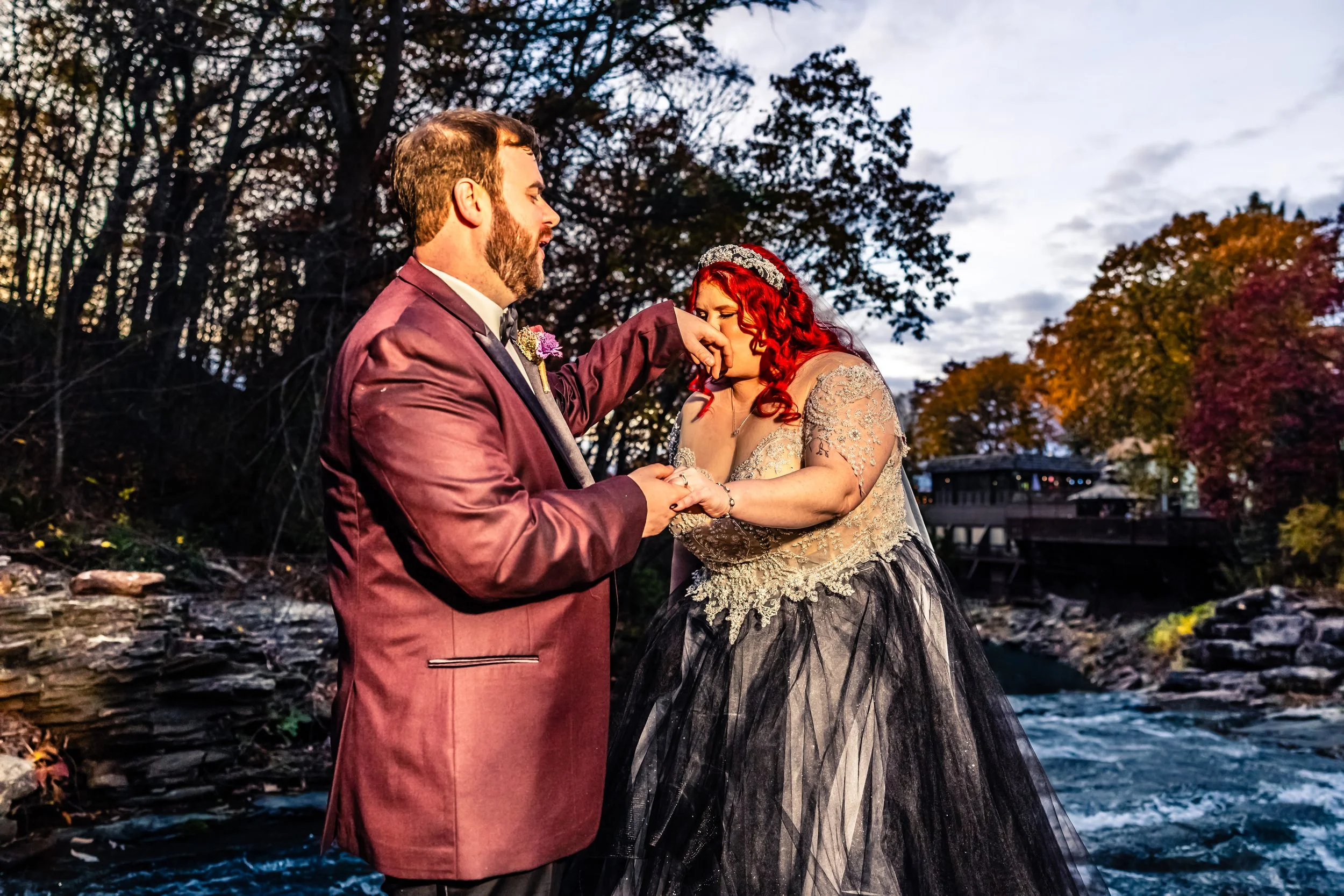 A newlywed couple stands by a river during sunset, with the groom kissing the bride's hand as she wipes tears of joy. The bride has red hair and wears a black and gold wedding gown, while the groom is in a burgundy tuxedo. Trees with autumn foliage a