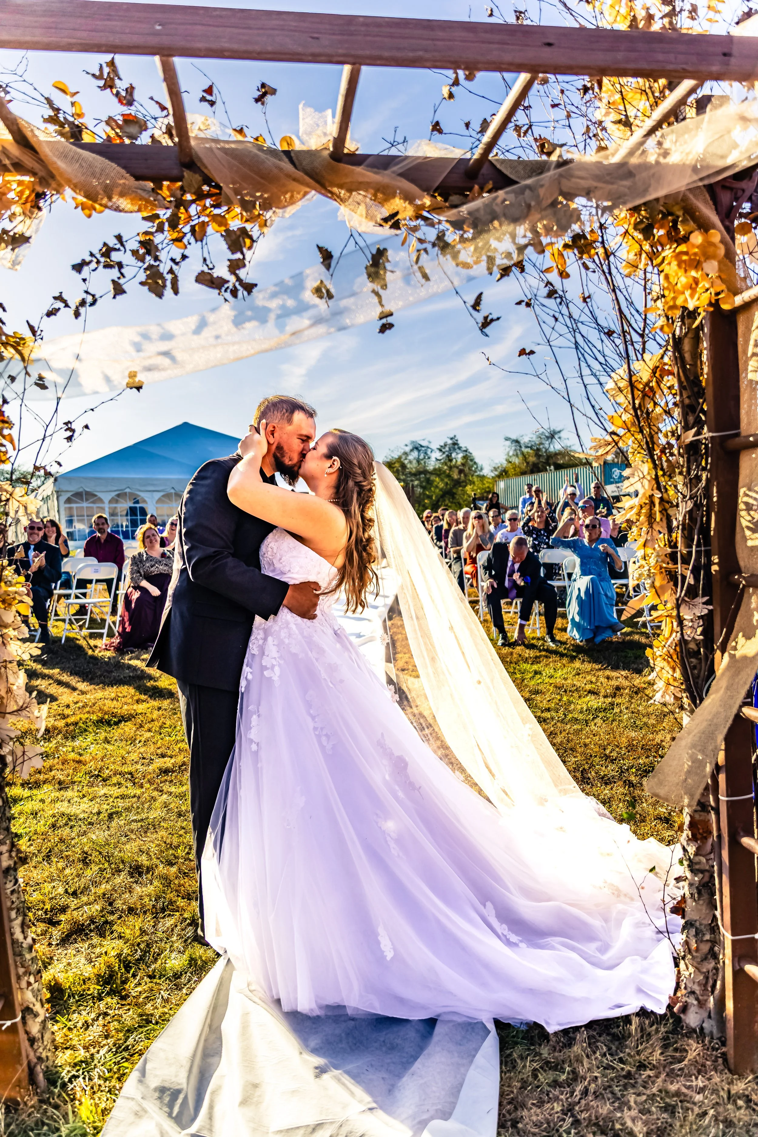A bride and groom kiss under a rustic wedding arch decorated with dried leaves and fabric, during their outdoor wedding ceremony in the late afternoon sunshine, with guests seated in the background.