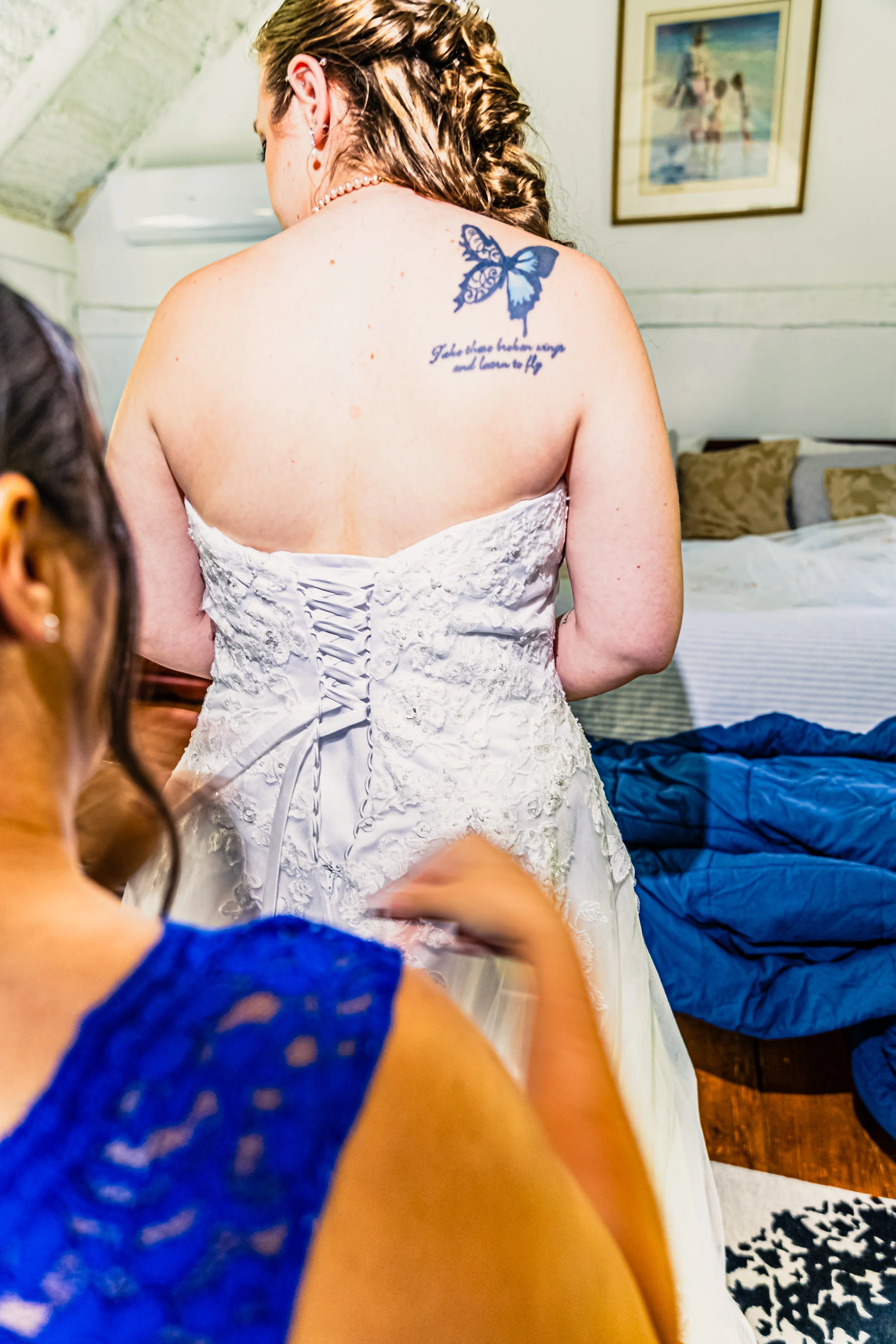 A bride in a strapless white wedding dress with lace details, getting ready for her wedding. A woman in a blue dress is helping her, and they are in a room with a bed and wall art.