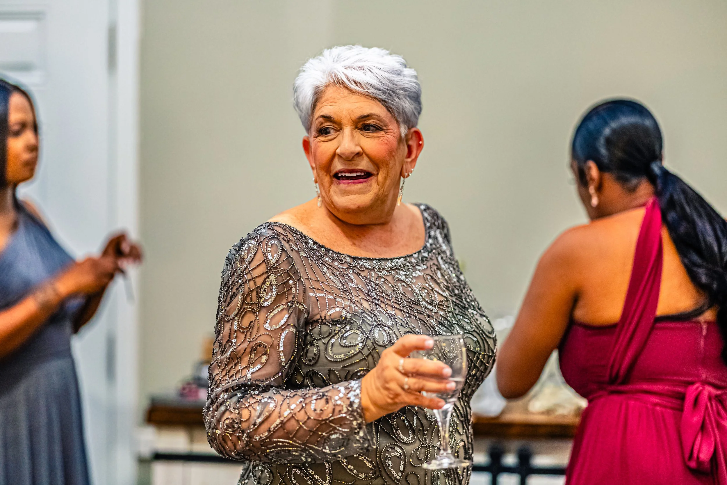 An elderly woman with short, white hair dressed in a sparkly silver dress holding a wine glass, talking to two women at a social gathering.