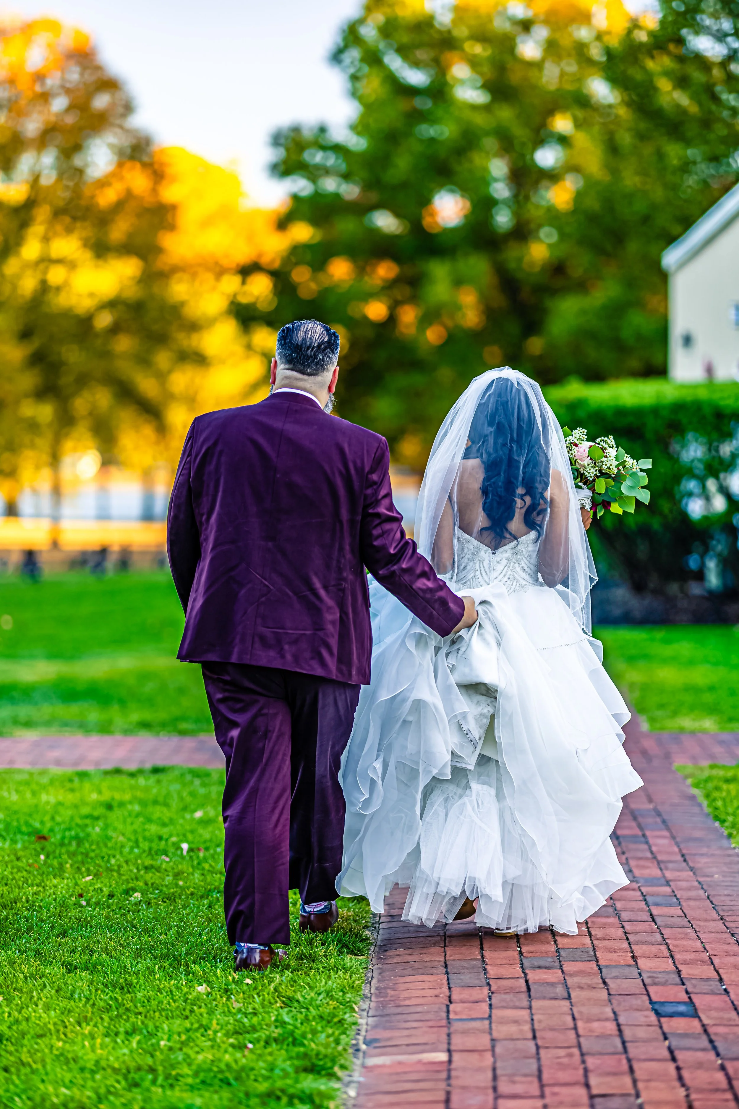 A bride and groom walking outdoors on a brick pathway during sunset, with the groom holding the bride's gown, and the bride carrying a bouquet of flowers.