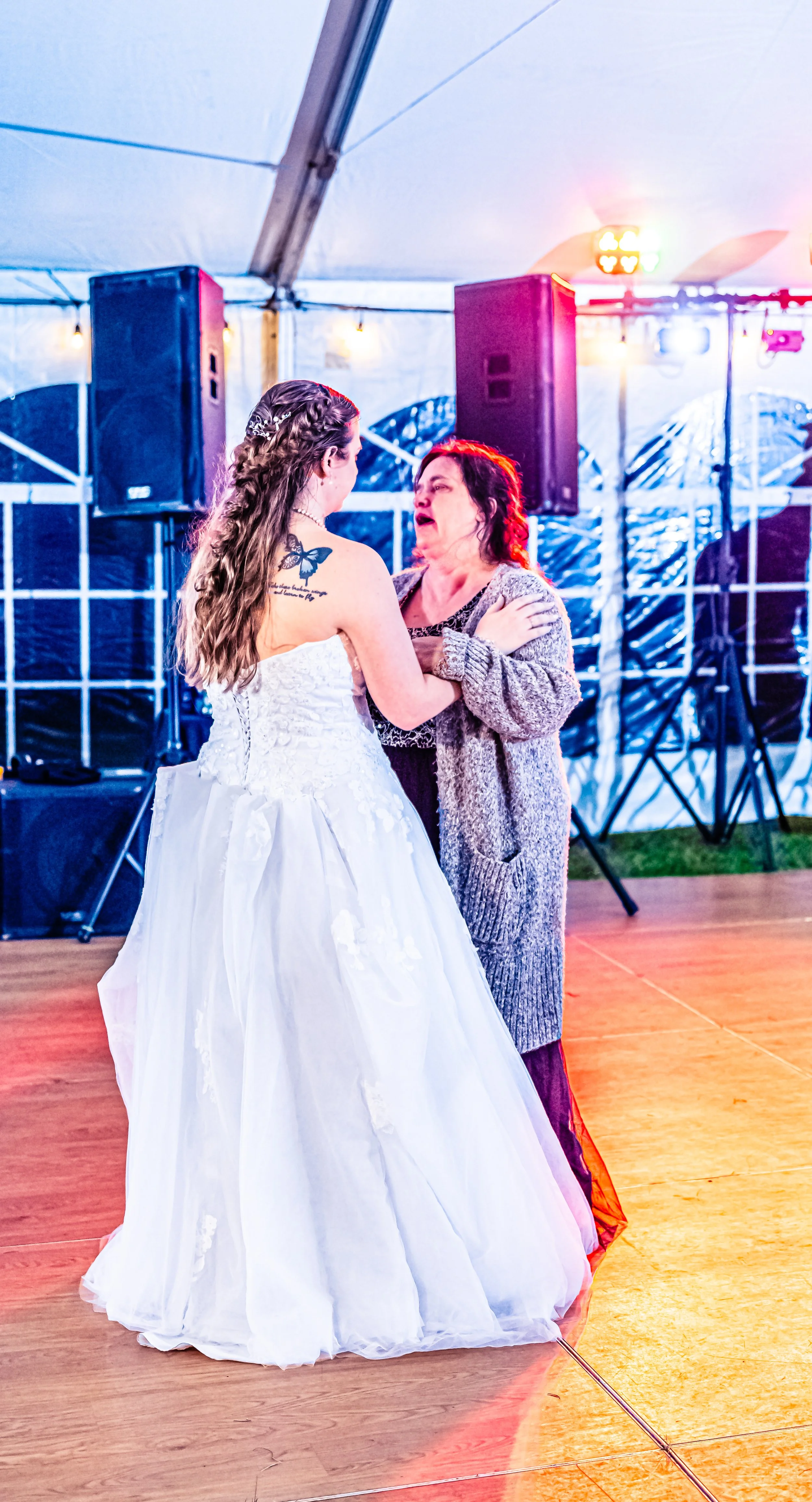 A bride in a white wedding gown dancing with a woman in a gray sweater in a tent with colorful lights and sound equipment in the background.