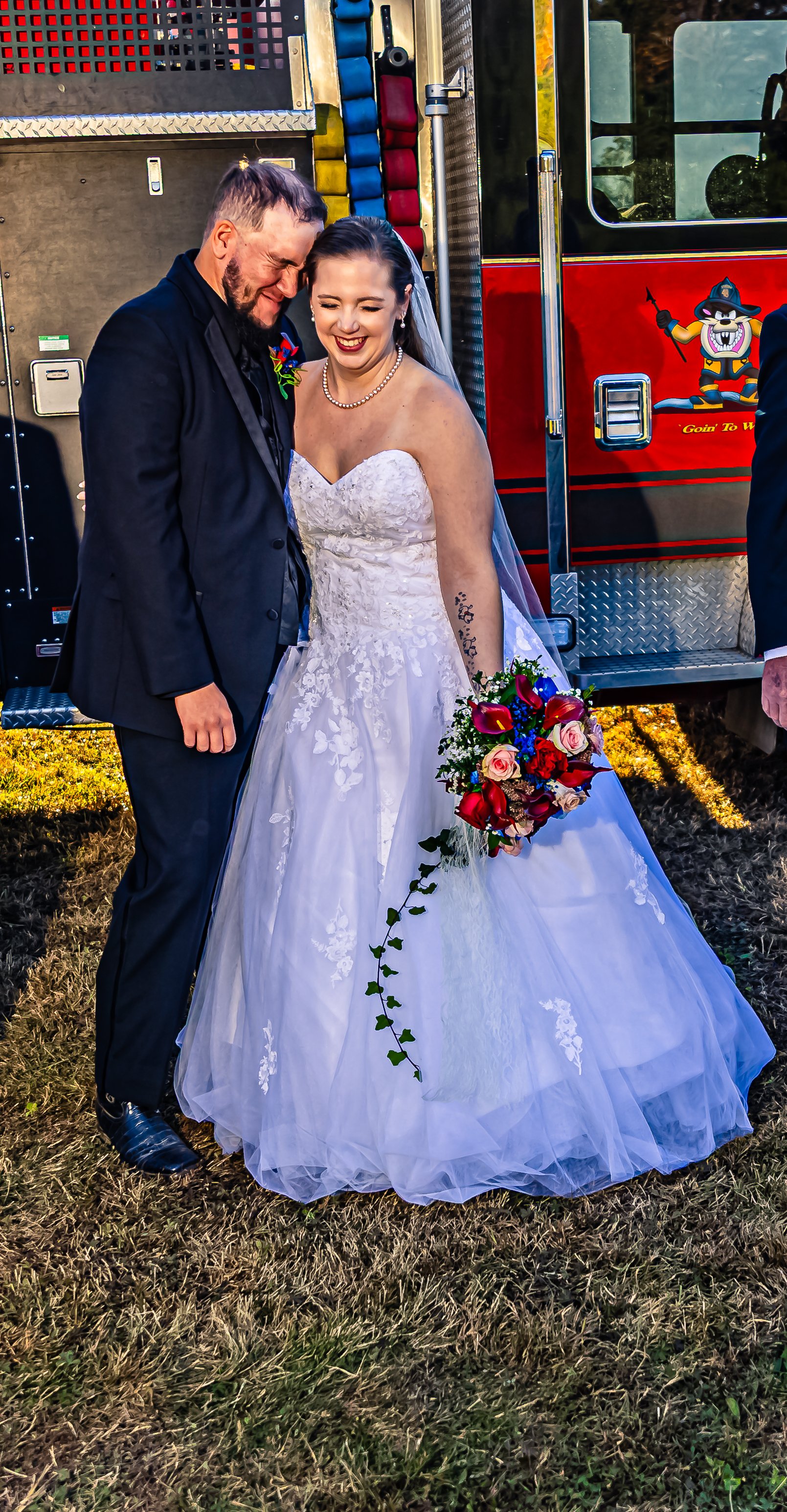 A bride and groom standing close together, smiling, in front of a fire truck, with the bride holding a bouquet of flowers.