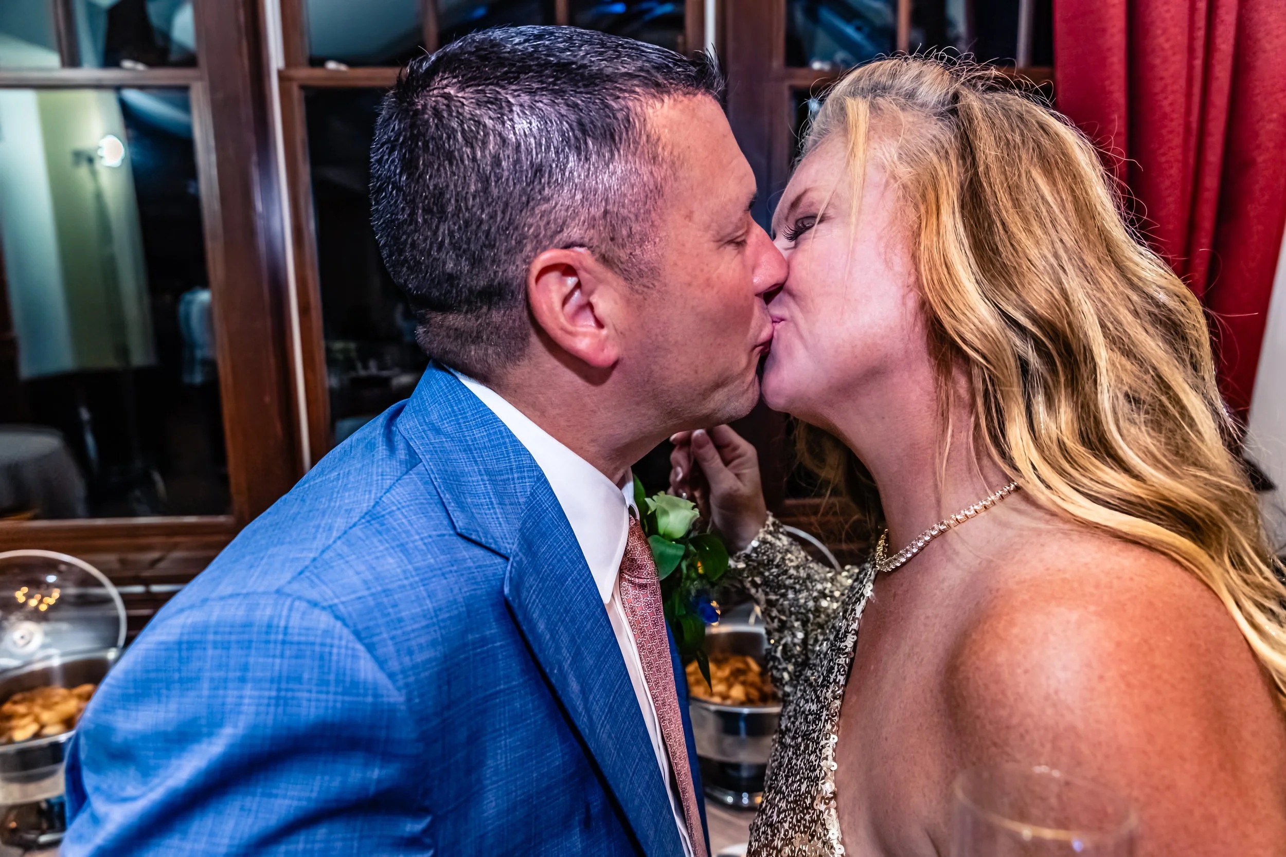 A man in a blue suit and a woman with long, wavy blonde hair and a sparkly dress kiss at a wedding reception