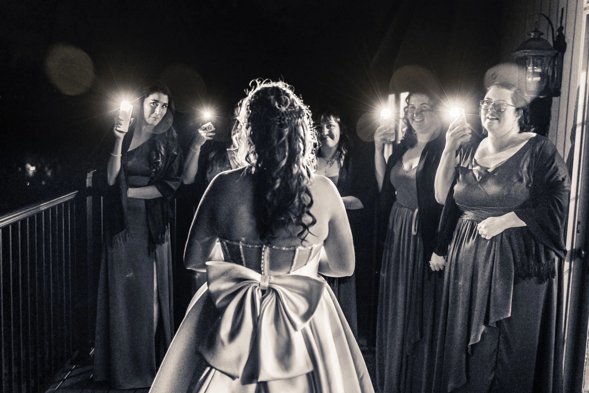Black and white photo showing a woman in a wedding dress facing five women who are holding up phones with flashlights.
