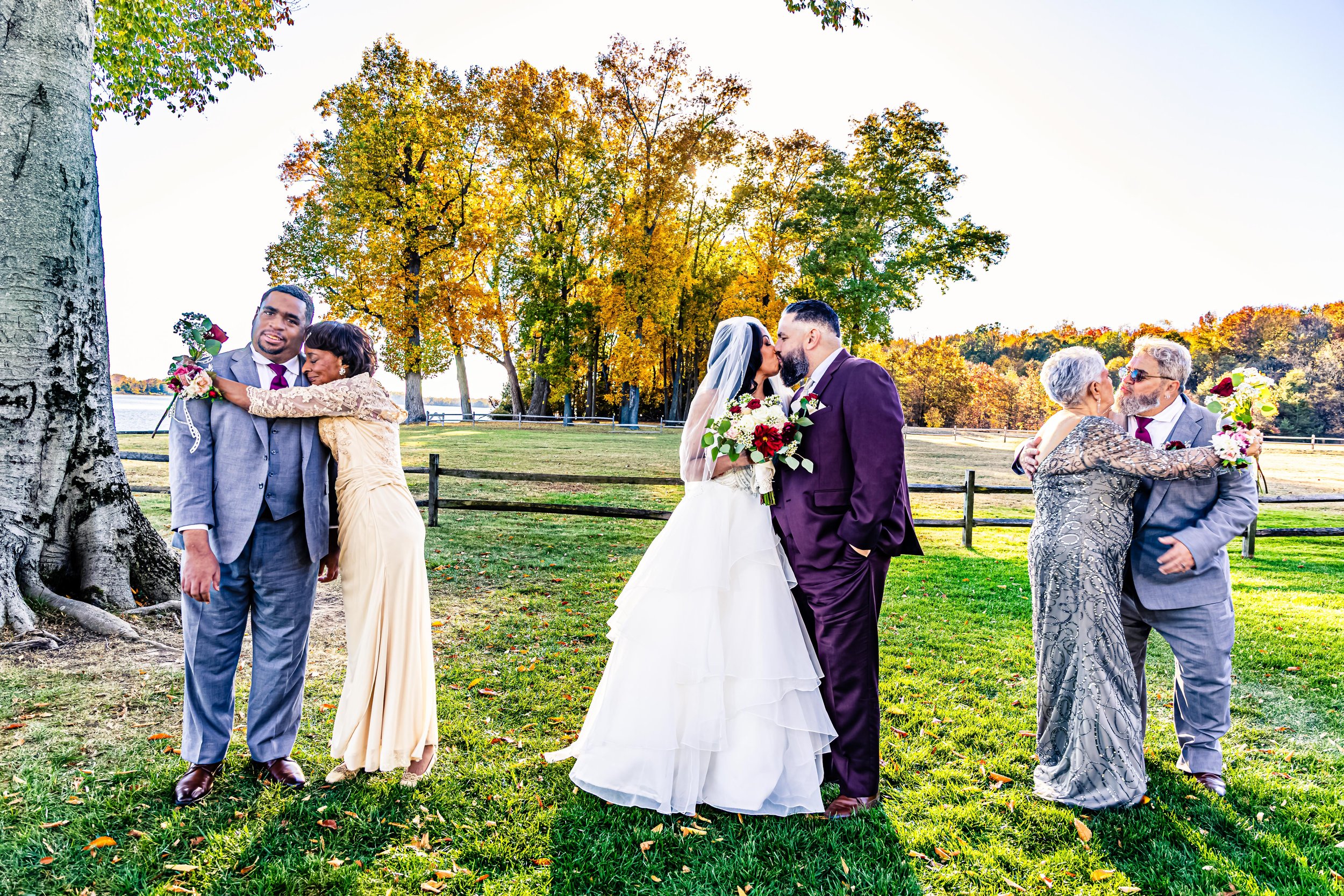 A wedding scene outdoors with two couples sharing affectionate moments, a man and woman kissing in the center, and two people hugging on each side. The setting features green grass and trees with autumn foliage.