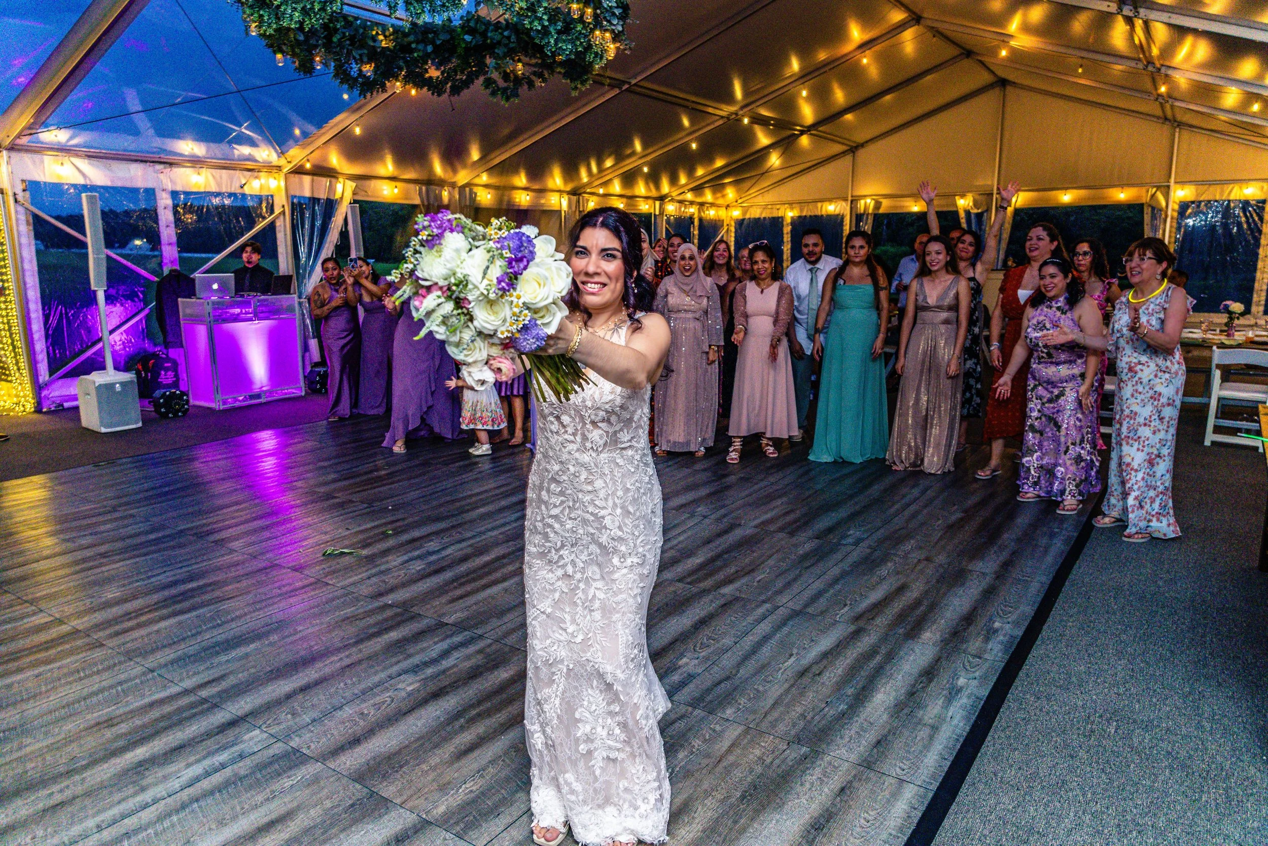 Bride in a lace wedding dress throws a bouquet of flowers during the wedding reception in a decorated tent with string lights, surrounded by guests in formal attire.